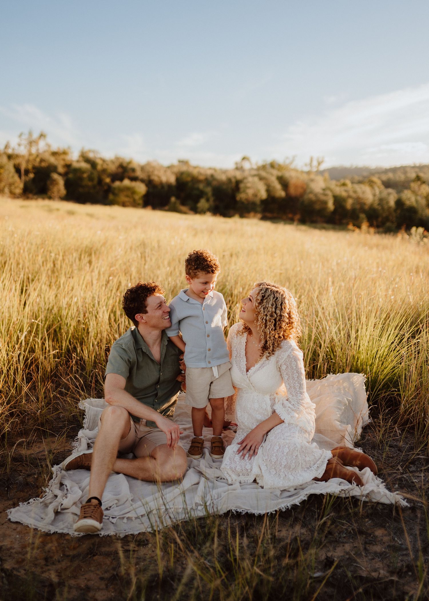 A family sits together on a white blanket in a golden field during sunset with trees in the background.