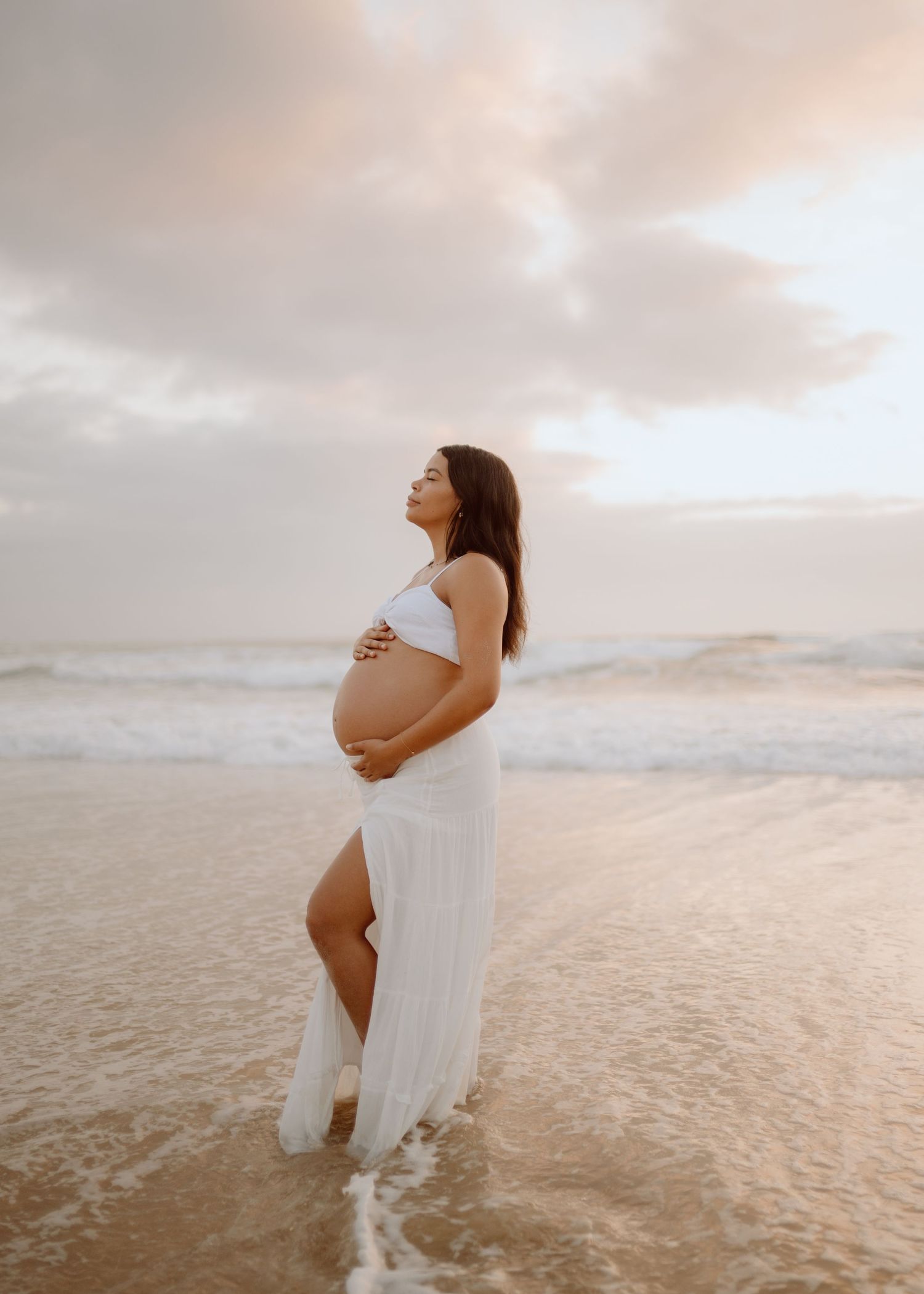 A serene beach maternity photoshoot at sunset shows a figure in a flowing white dress standing in shallow ocean water.