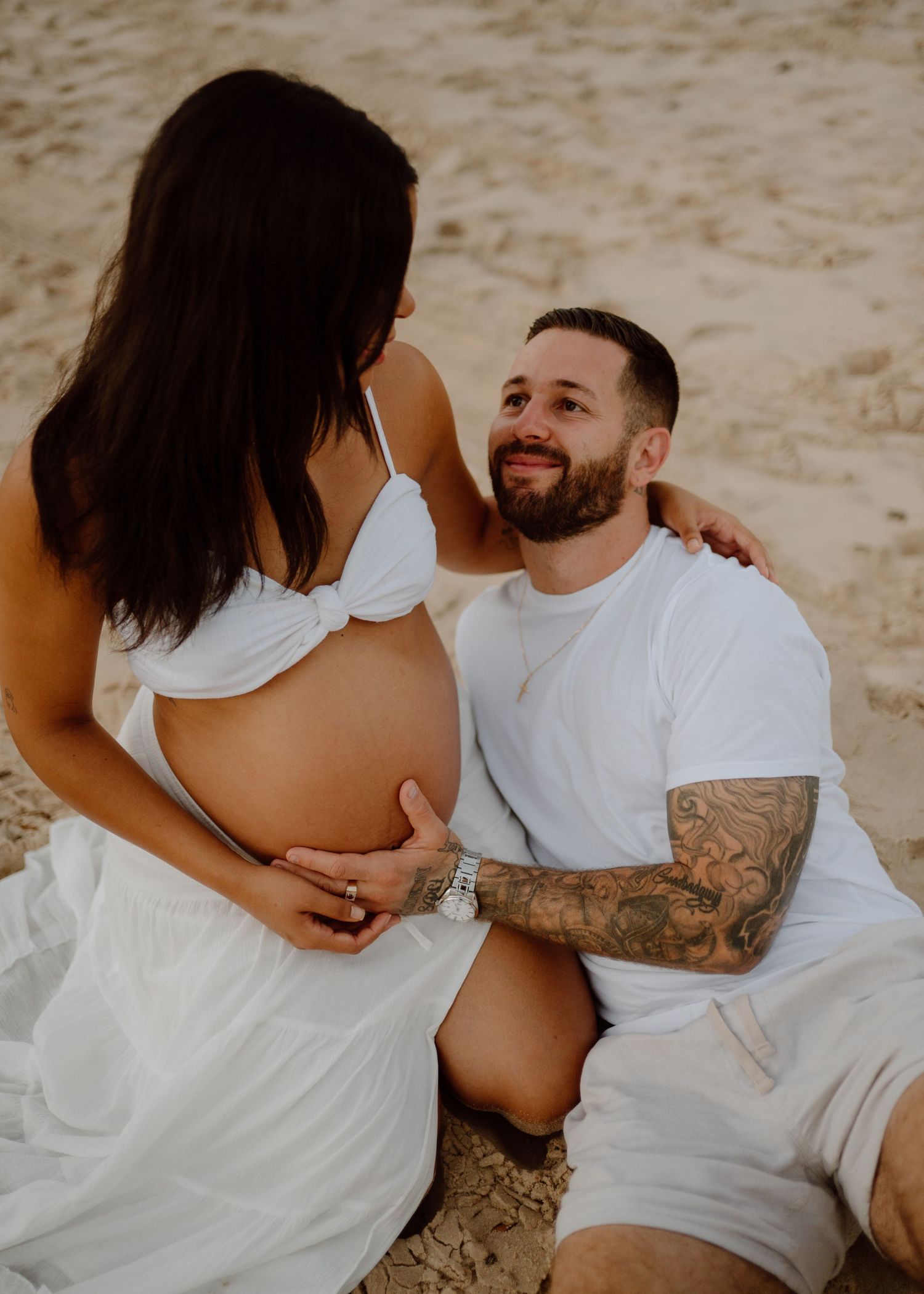 A series of intimate maternity photos showing a couple embracing on a sandy beach in white clothing.