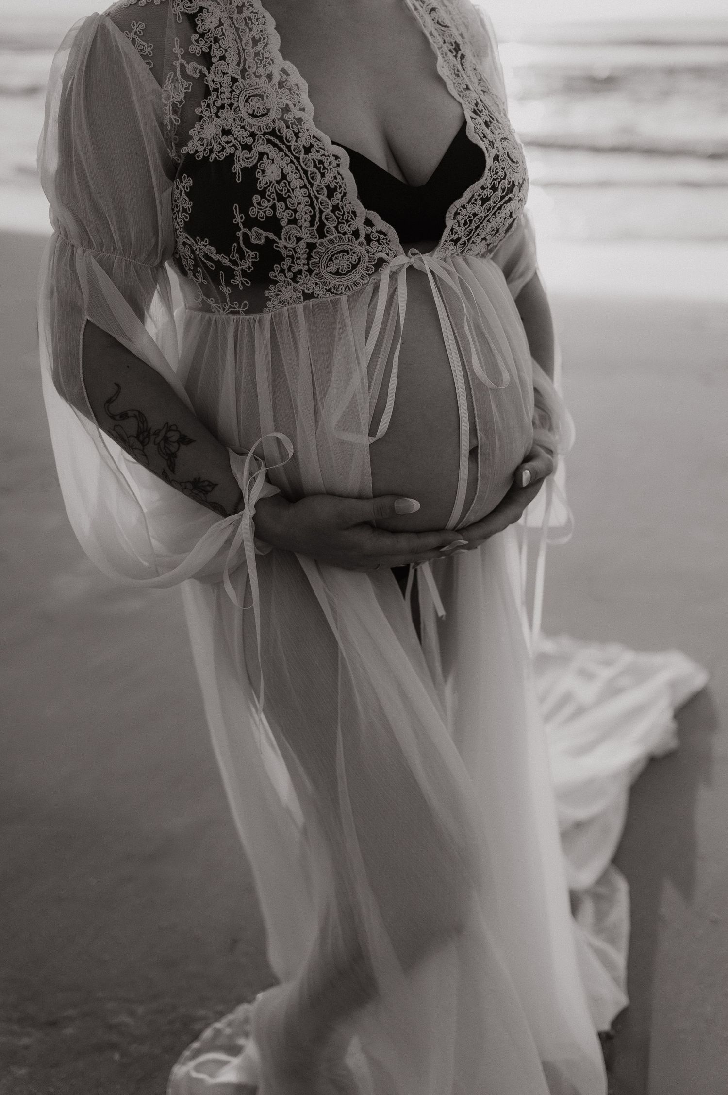 Black and white maternity photo of pregnant woman in lace robe cradling baby bump on beach.