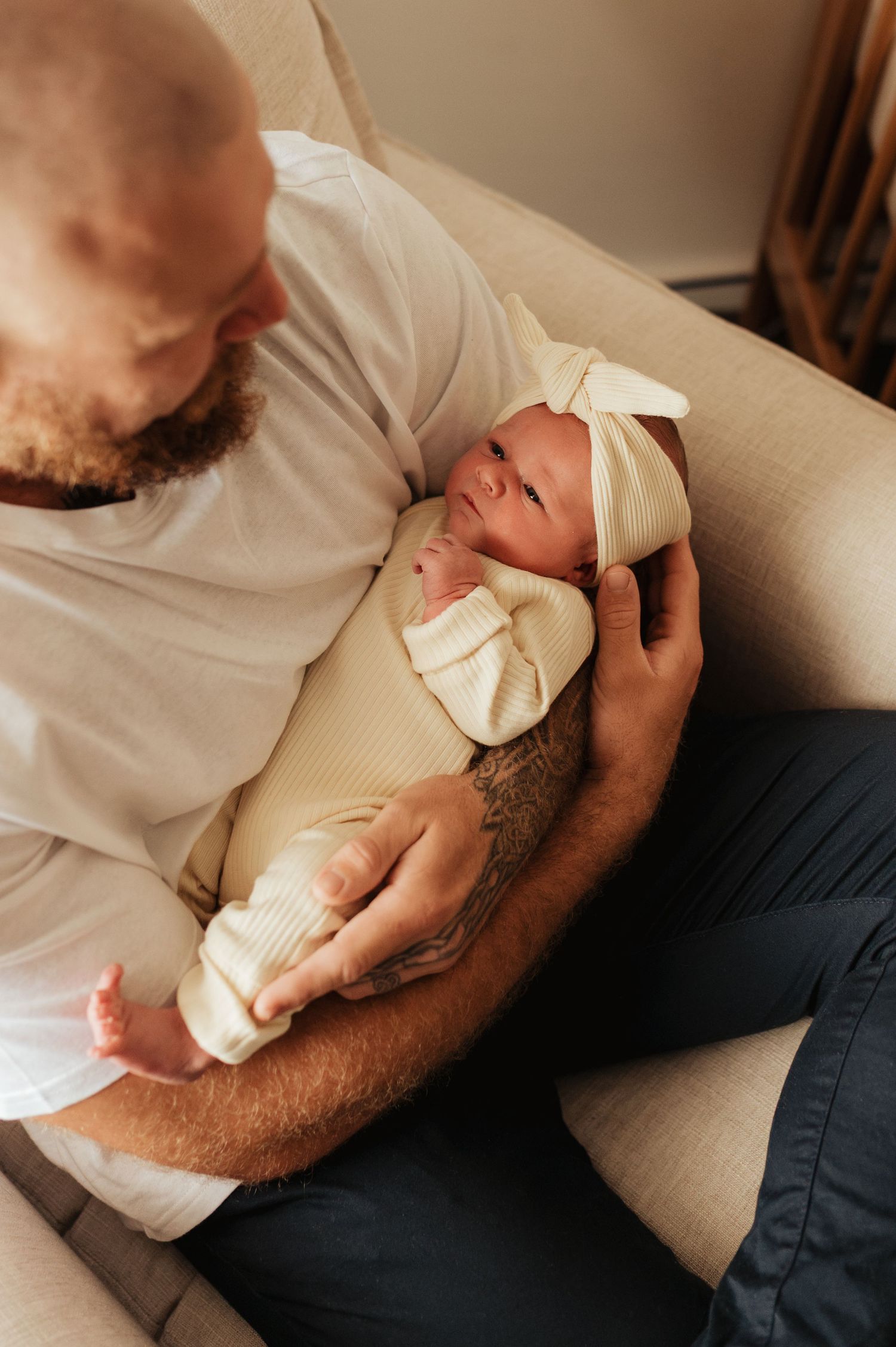 Bald bearded father with tattoos cradling sleeping newborn baby in cream swaddle and bow hat on chair.