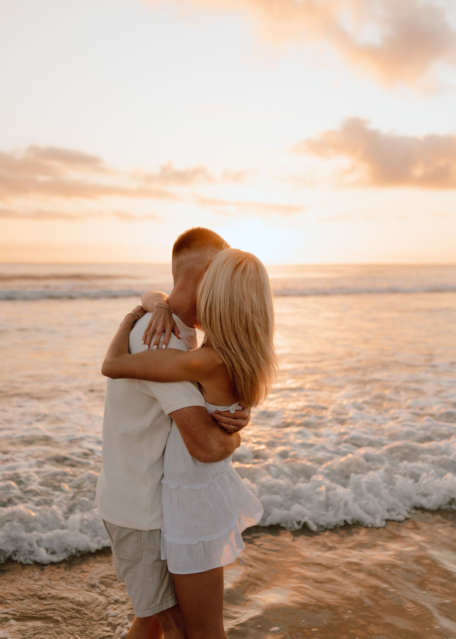 A couple embraces on a sandy beach during golden hour sunset as ocean waves roll in behind them.