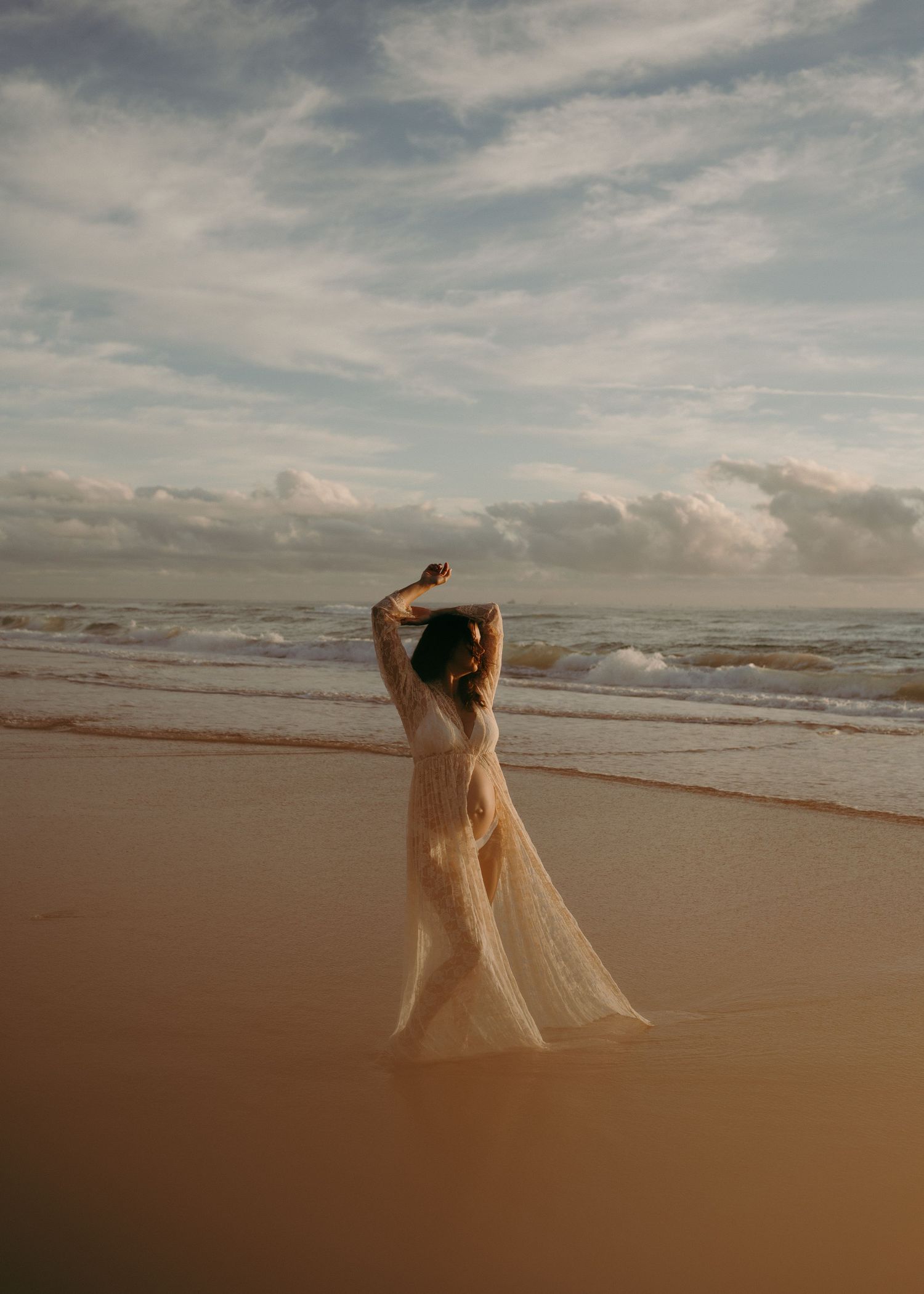 A graceful figure in a flowing dress poses elegantly on a sandy beach at sunset with waves and cloudy skies in the background.