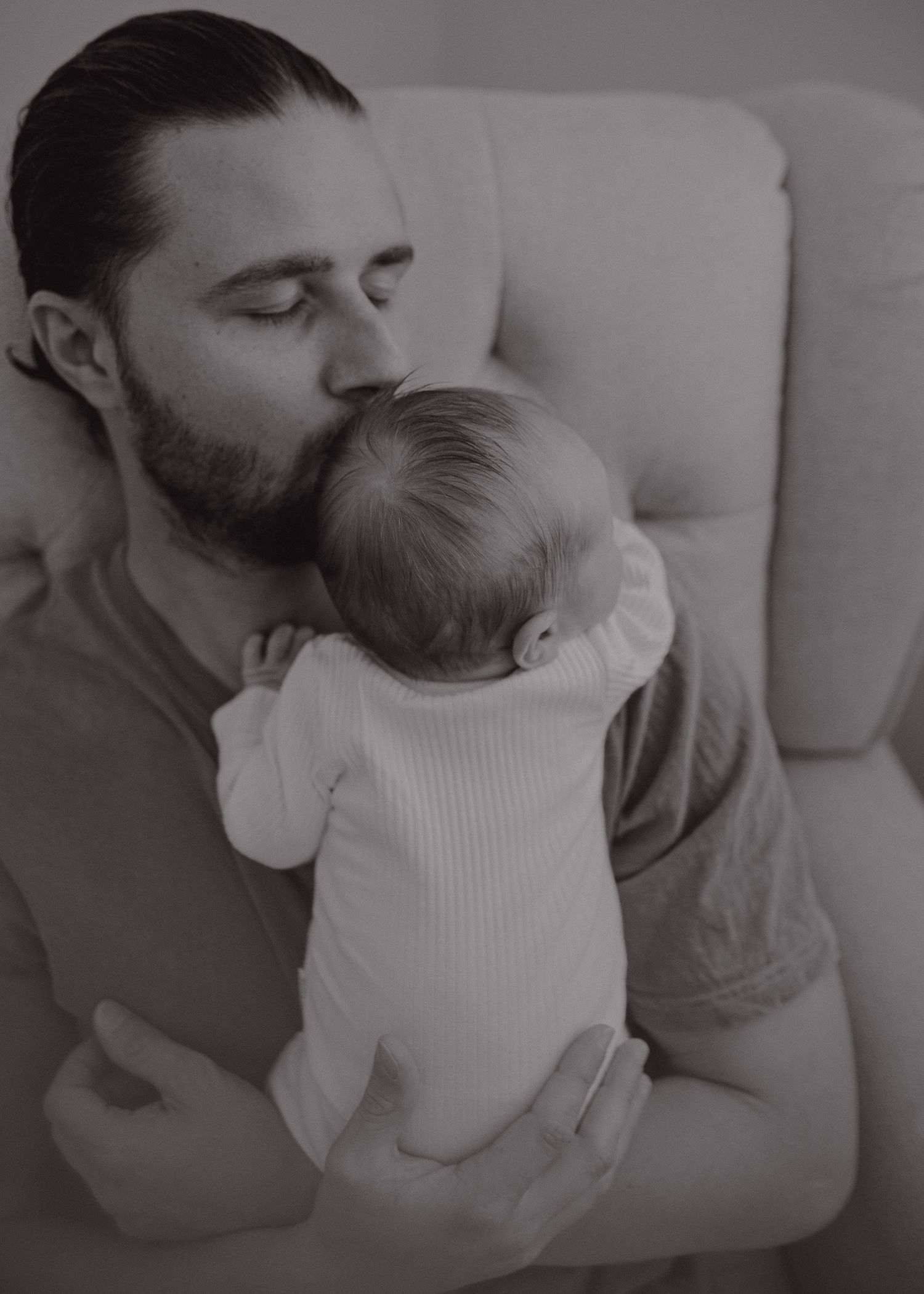 A tender black and white photo of a parent holding and kissing a baby in a white onesie while resting on a couch.