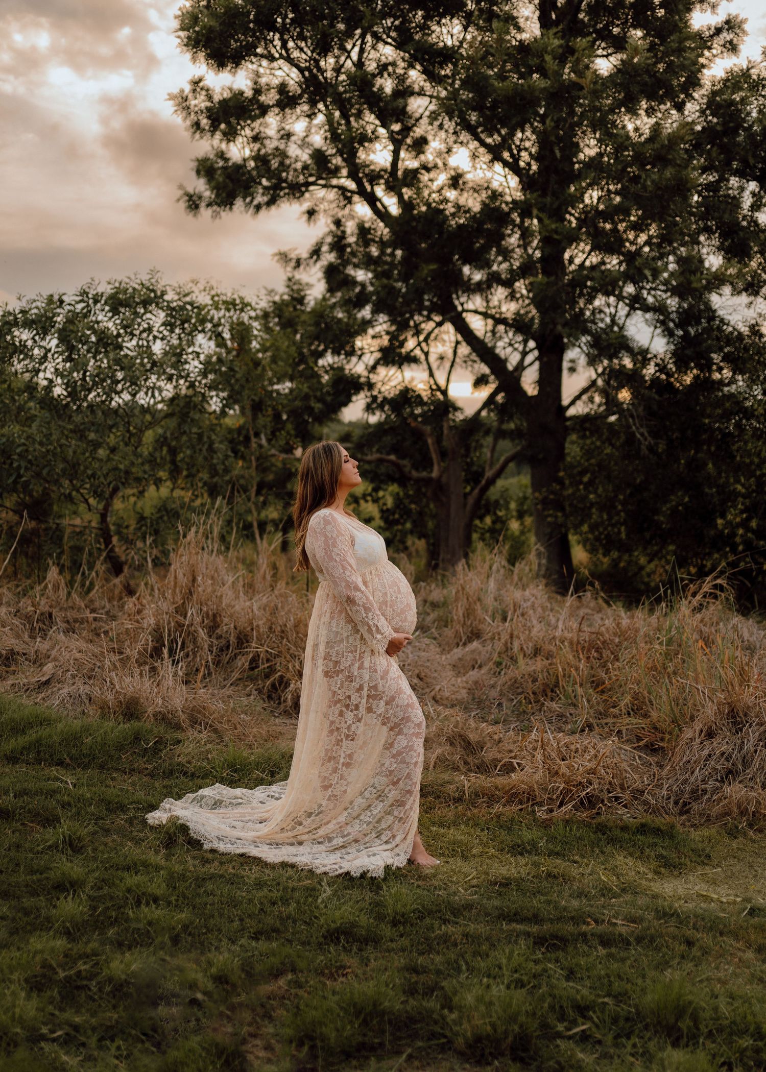 A person in a flowing lace dress stands in a field at sunset with tall trees silhouetted in the background.
