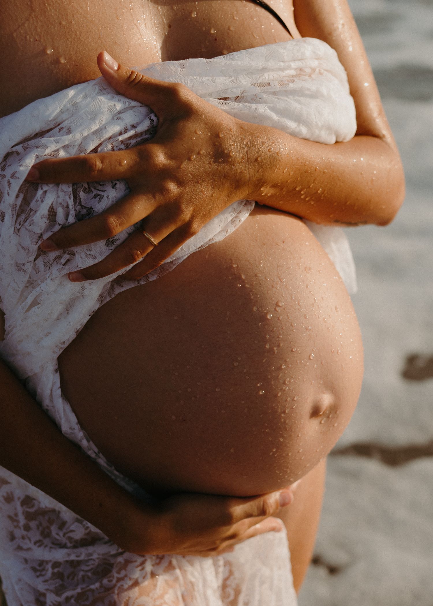 A pregnant woman's belly is shown on a beach with waves crashing in the background and white foam on her skin.