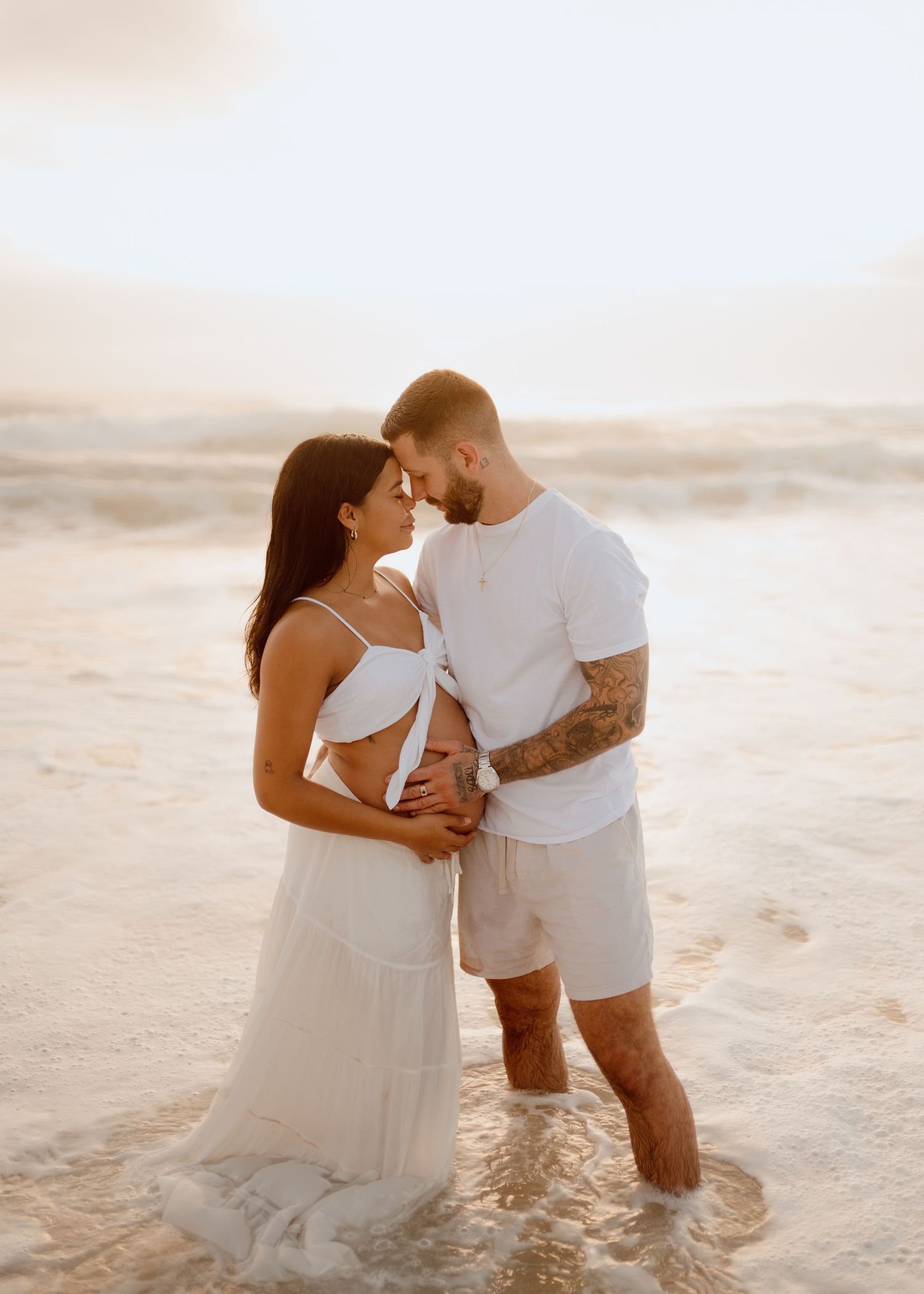 Couple in white outfits share intimate moments during a romantic beach photoshoot at sunset.