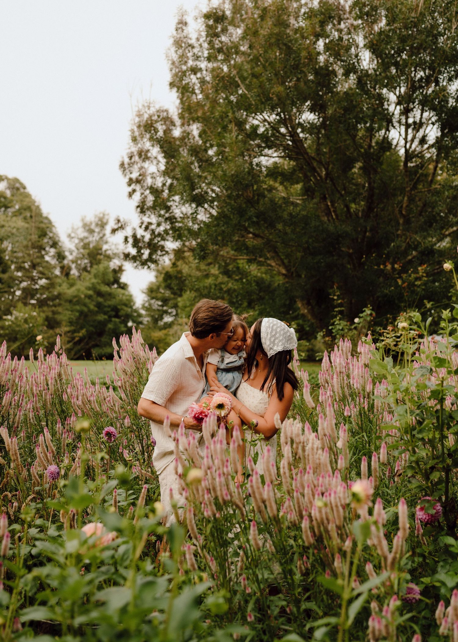A couple sharing a tender moment with their young child among pink wildflowers in a garden setting.