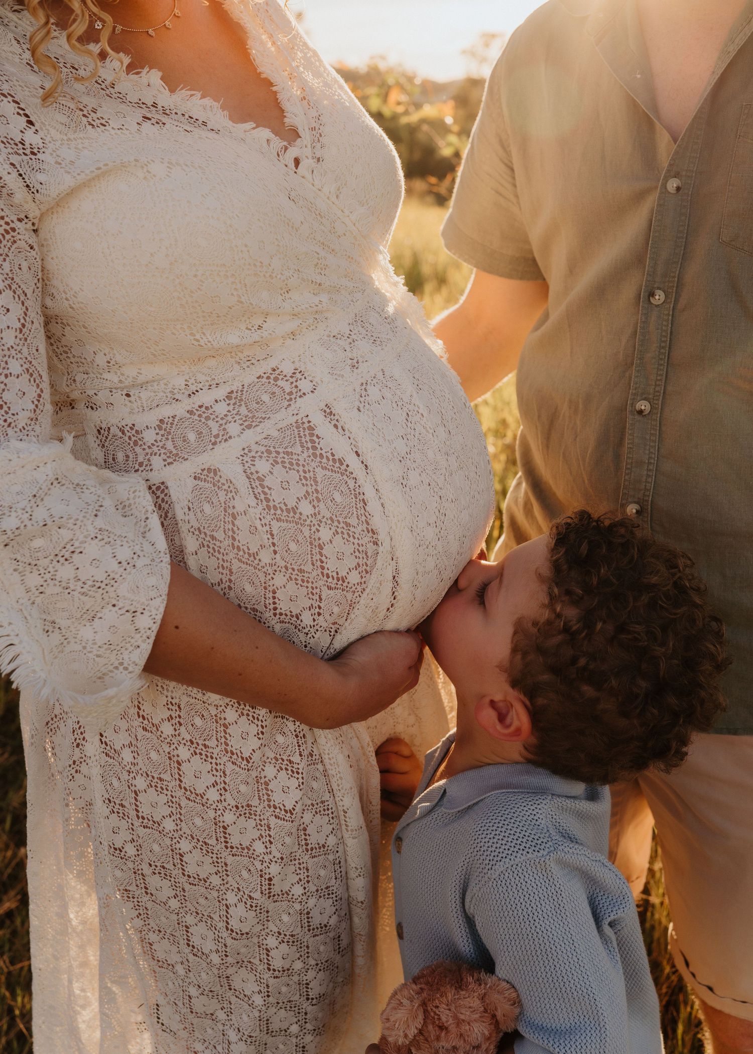 A couple embracing and kissing outdoors with sunlight filtering through, bride wearing delicate white lace.