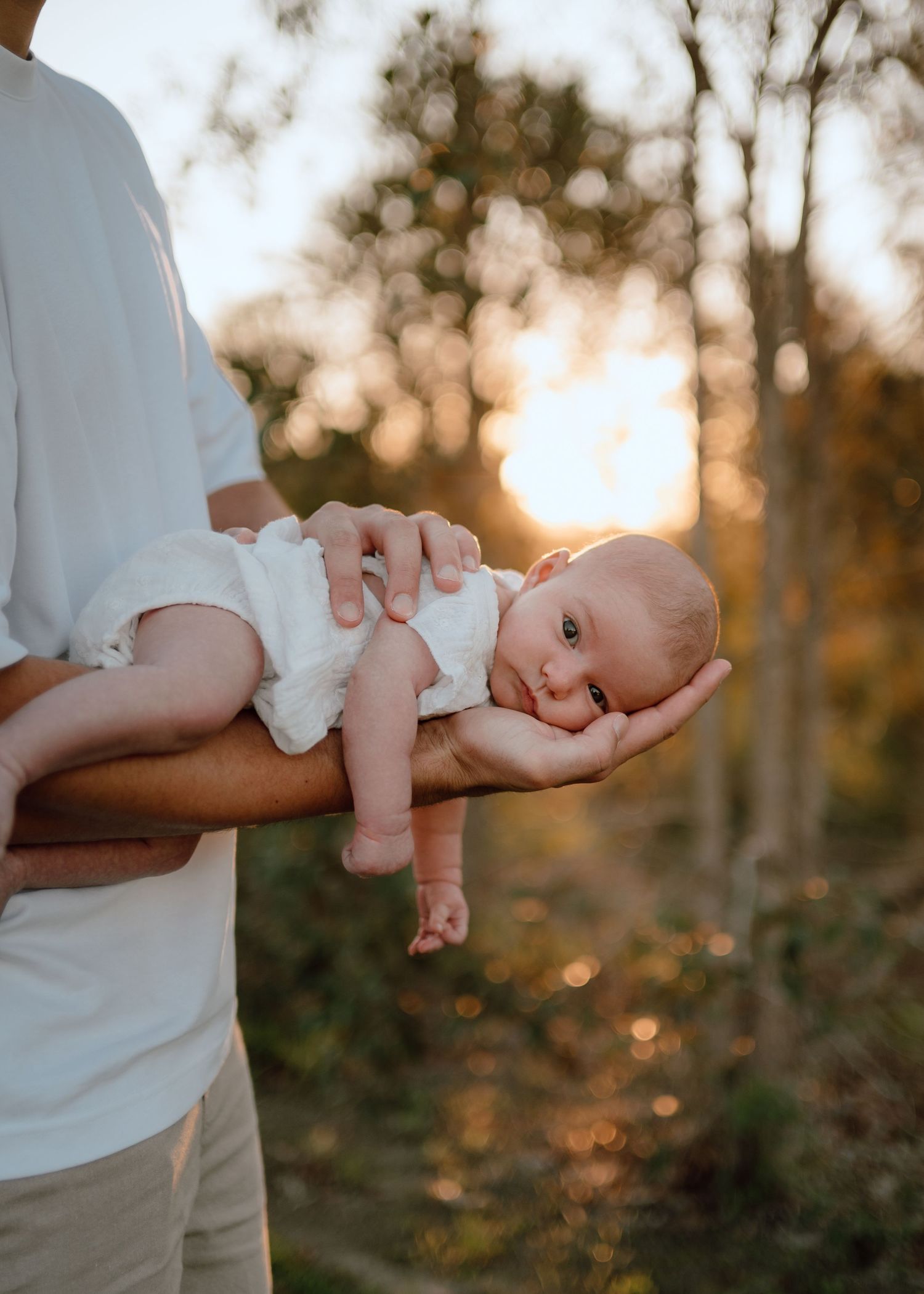A baby in white clothing is held during a sunset outdoor photography session.