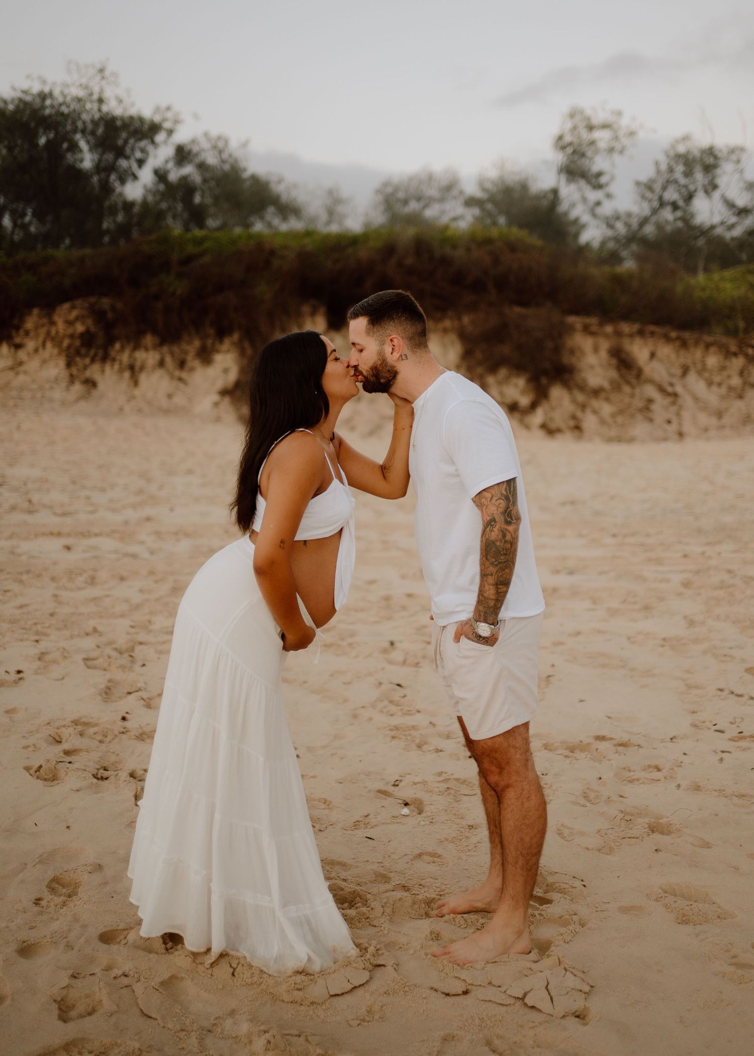 A couple shares a romantic moment in white outfits on a sandy beach during sunset.