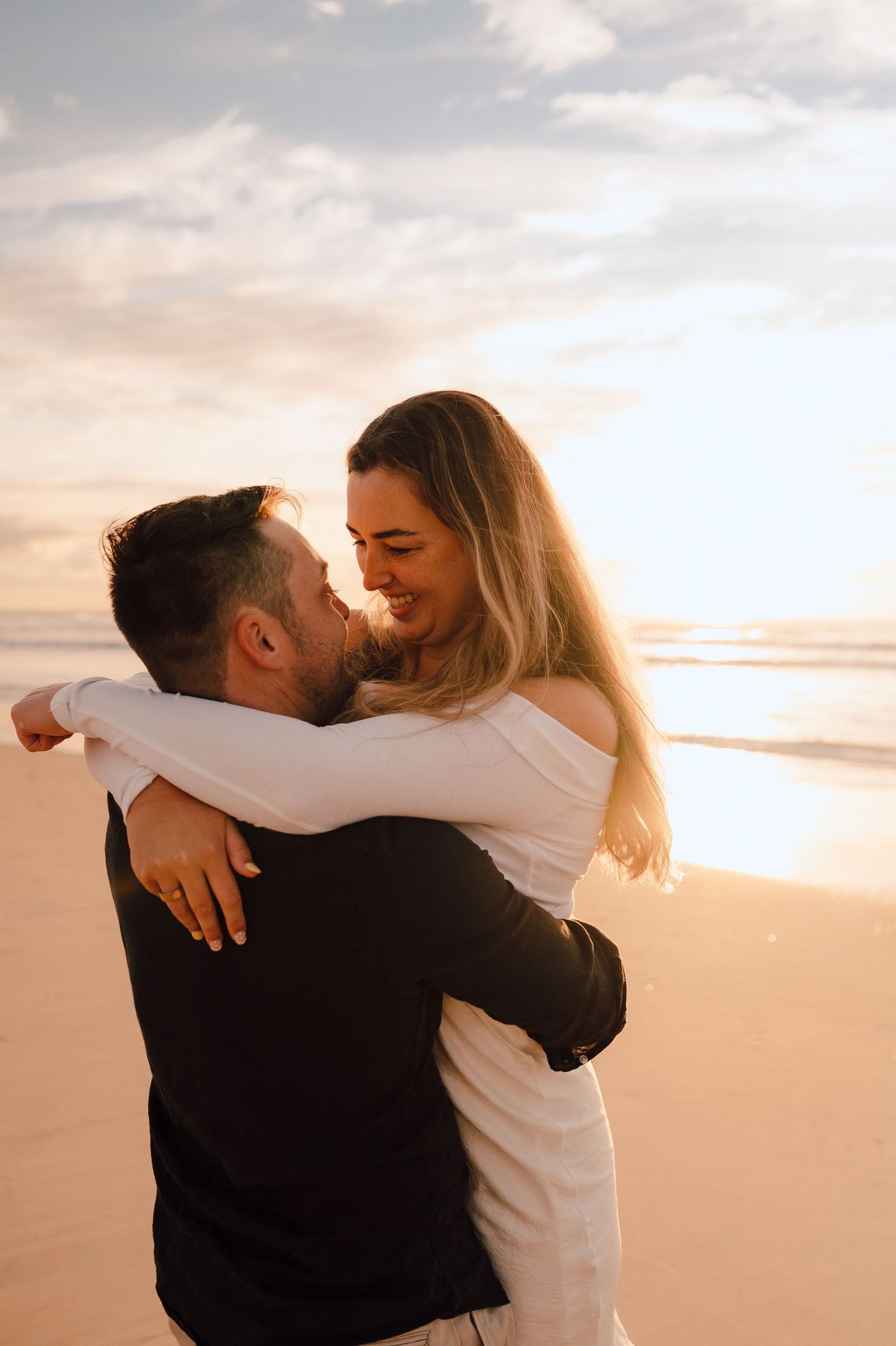 Couple embracing on a sandy beach at golden sunset, woman in white dress smiling joyfully.