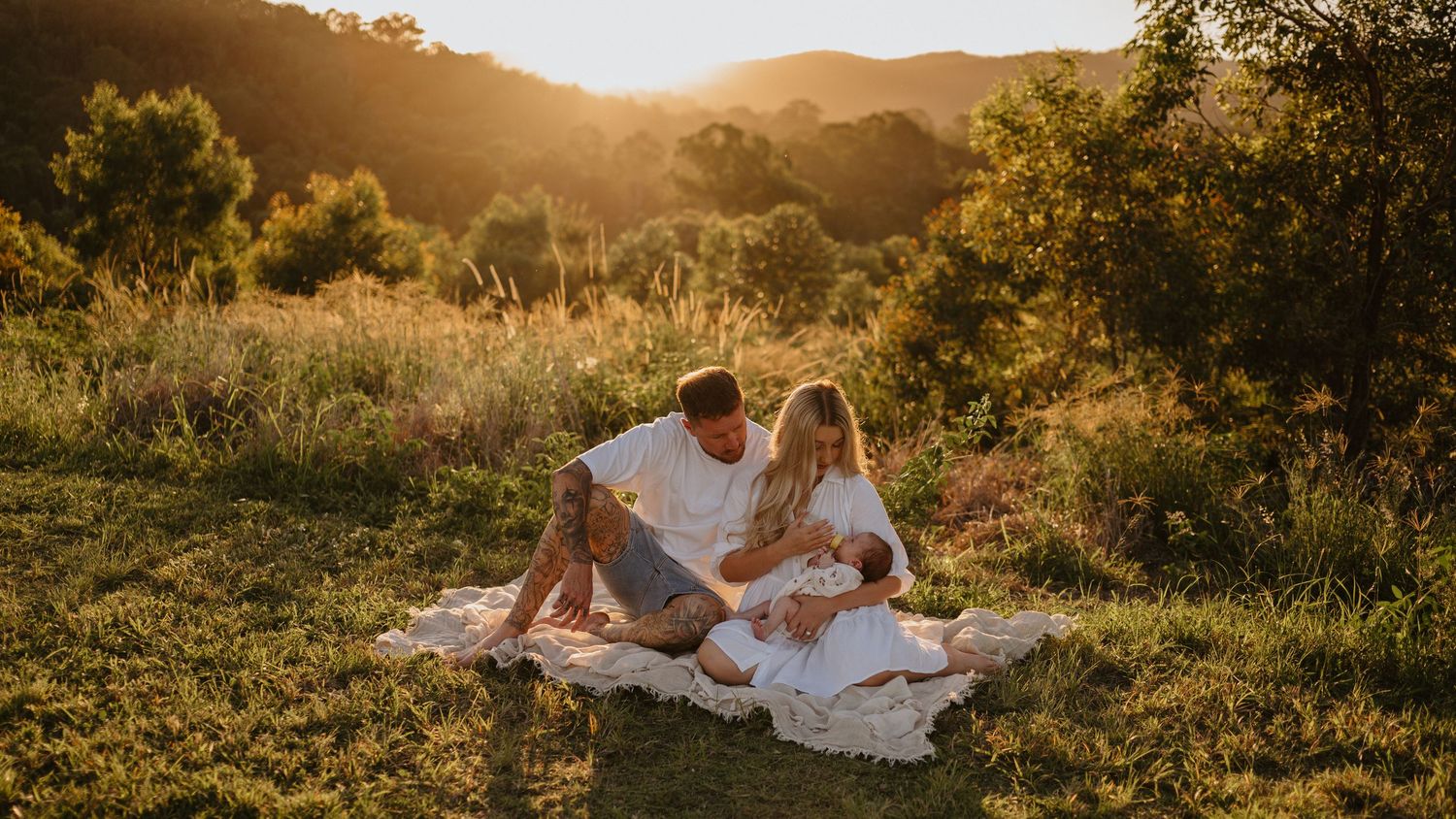 Family sitting on blanket in golden sunset meadow with lush green hills, parents gazing lovingly at newborn baby.