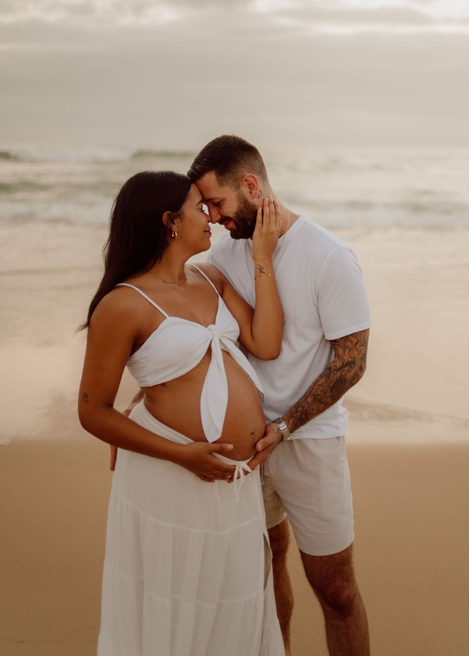 A romantic couple in white outfits shares intimate moments during a sunset beach photoshoot.