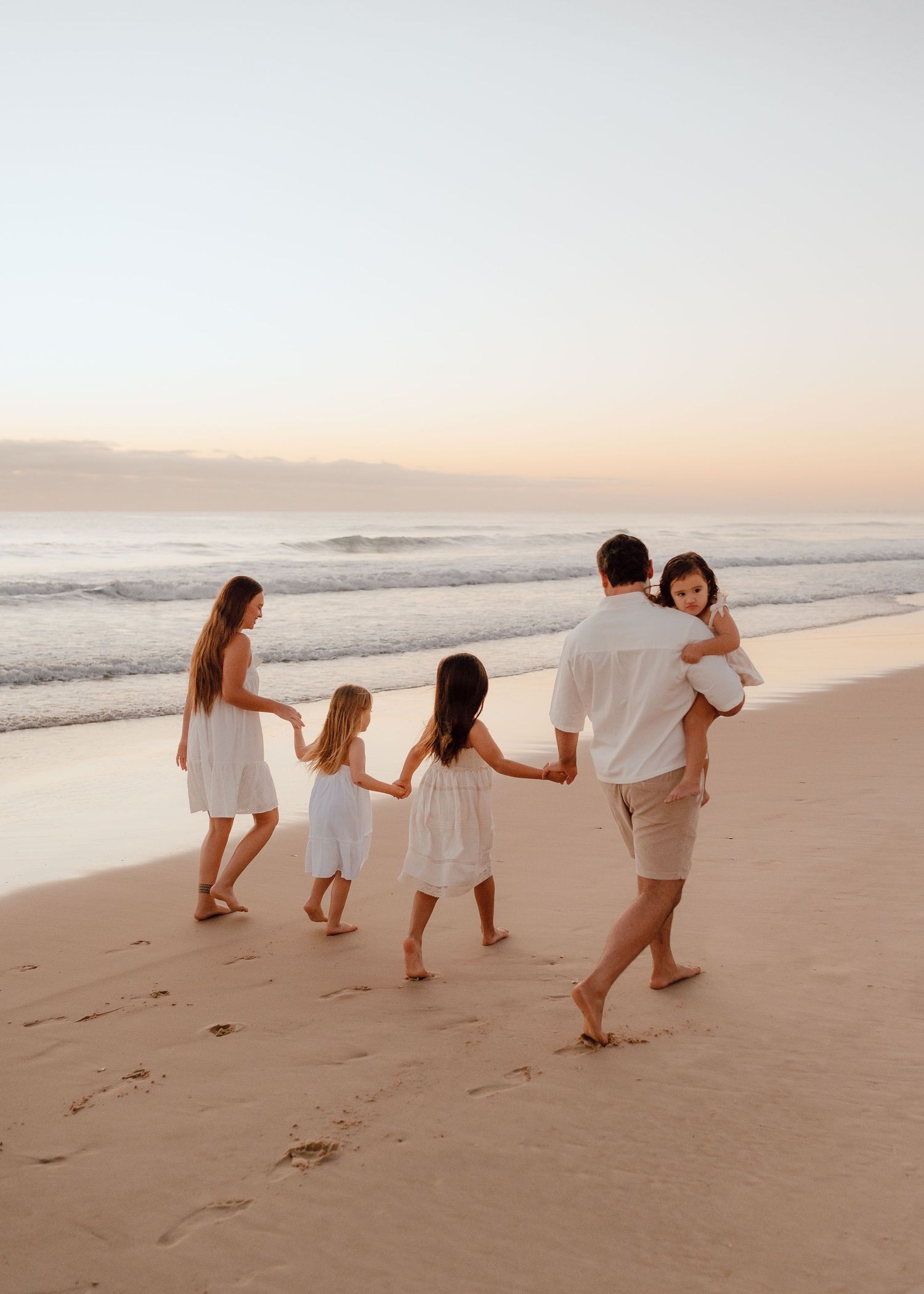 A family wearing white outfits playfully poses together on a sandy beach at sunset.