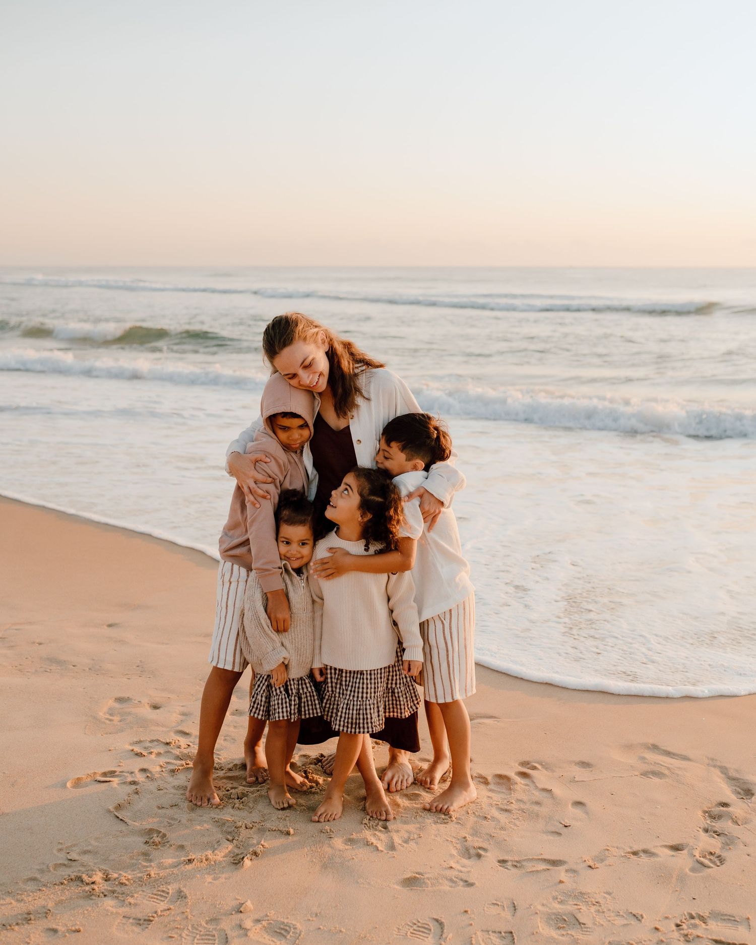 Sunset beach portrait with group hugging in coordinated neutral outfits.