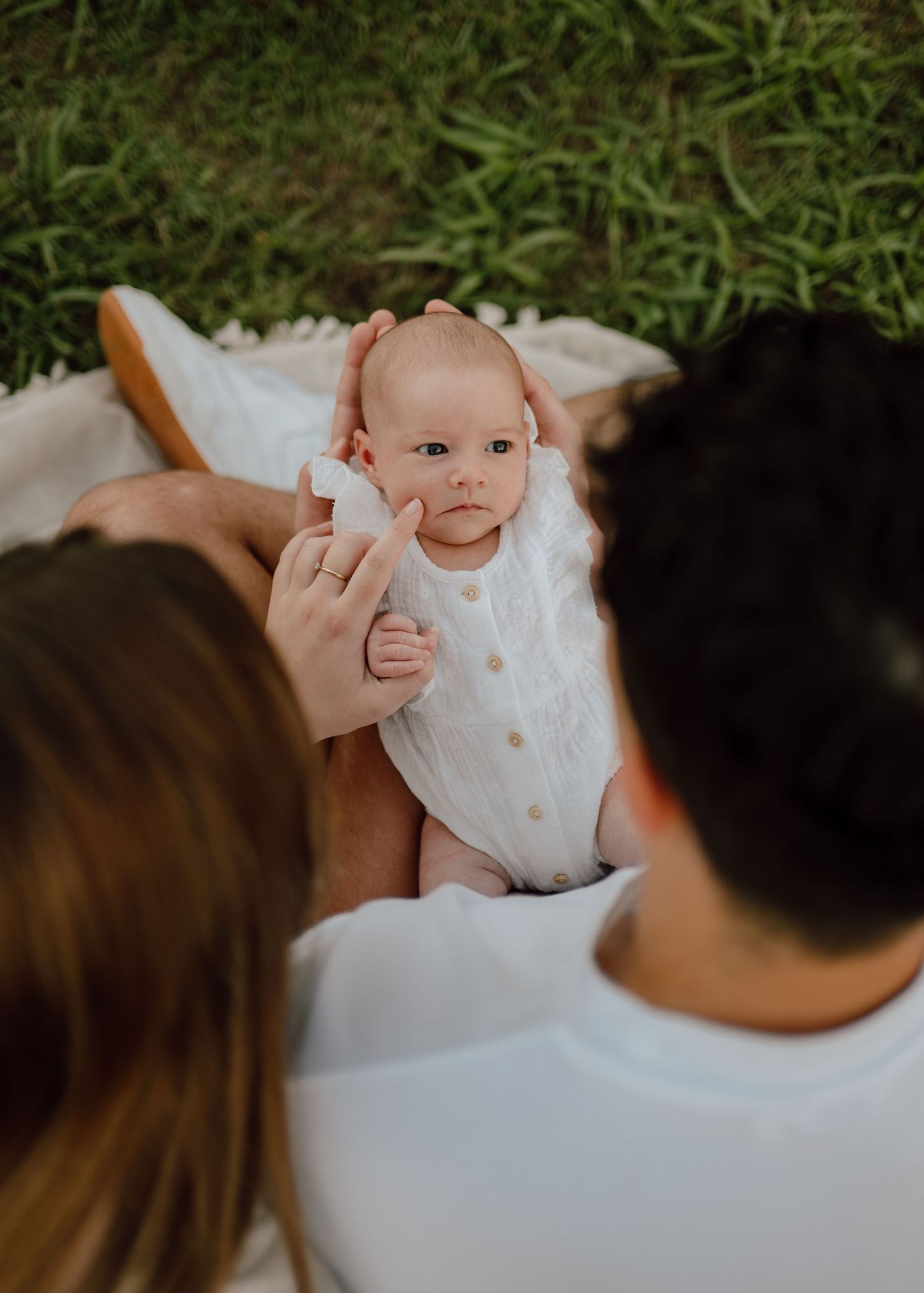 A baby wearing white clothing smiles during an outdoor family photoshoot.