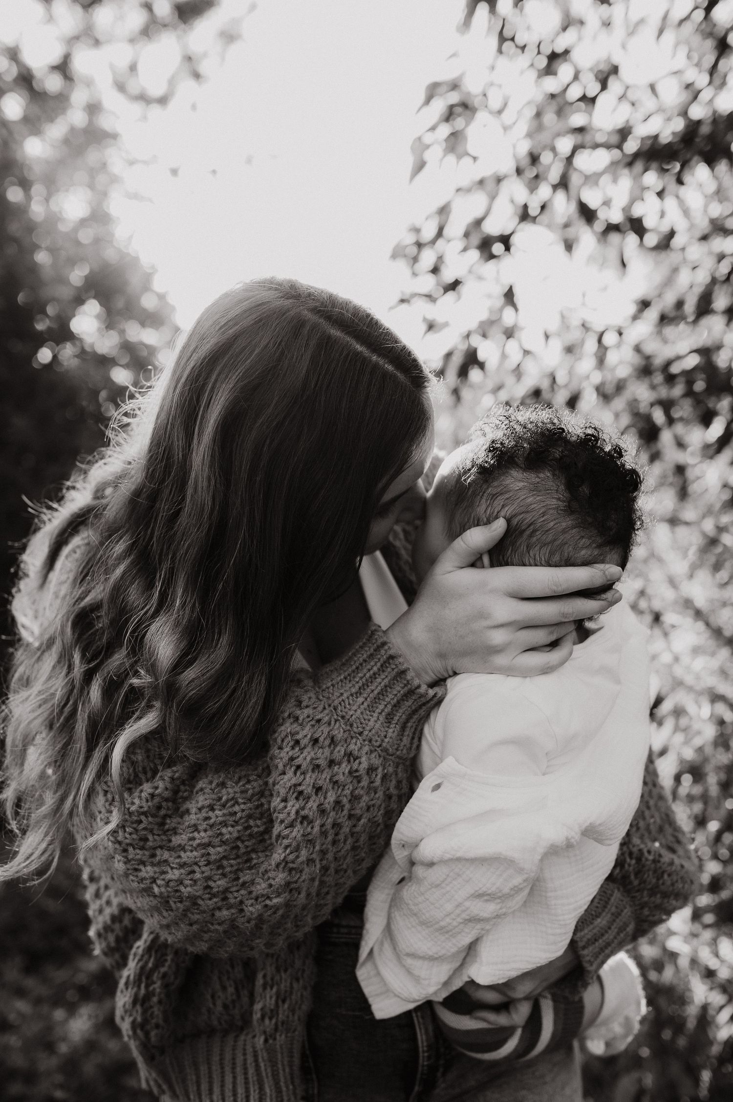 Black and white photo of a mother tenderly kissing her newborn baby outdoors among lush trees.