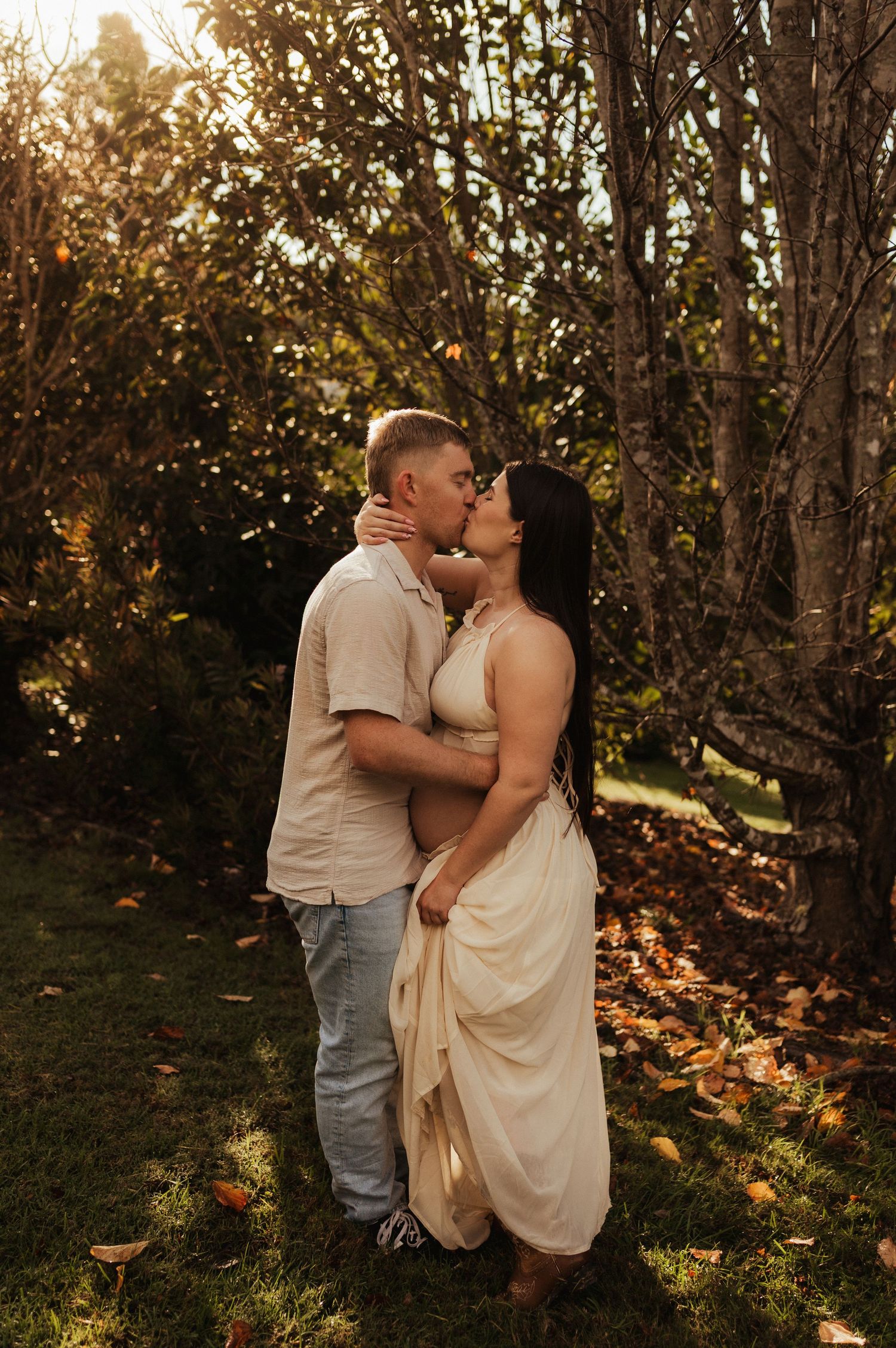 Couple sharing a romantic kiss in golden hour sunlight, woman in cream dress with visible baby bump, surrounded by autumn trees.