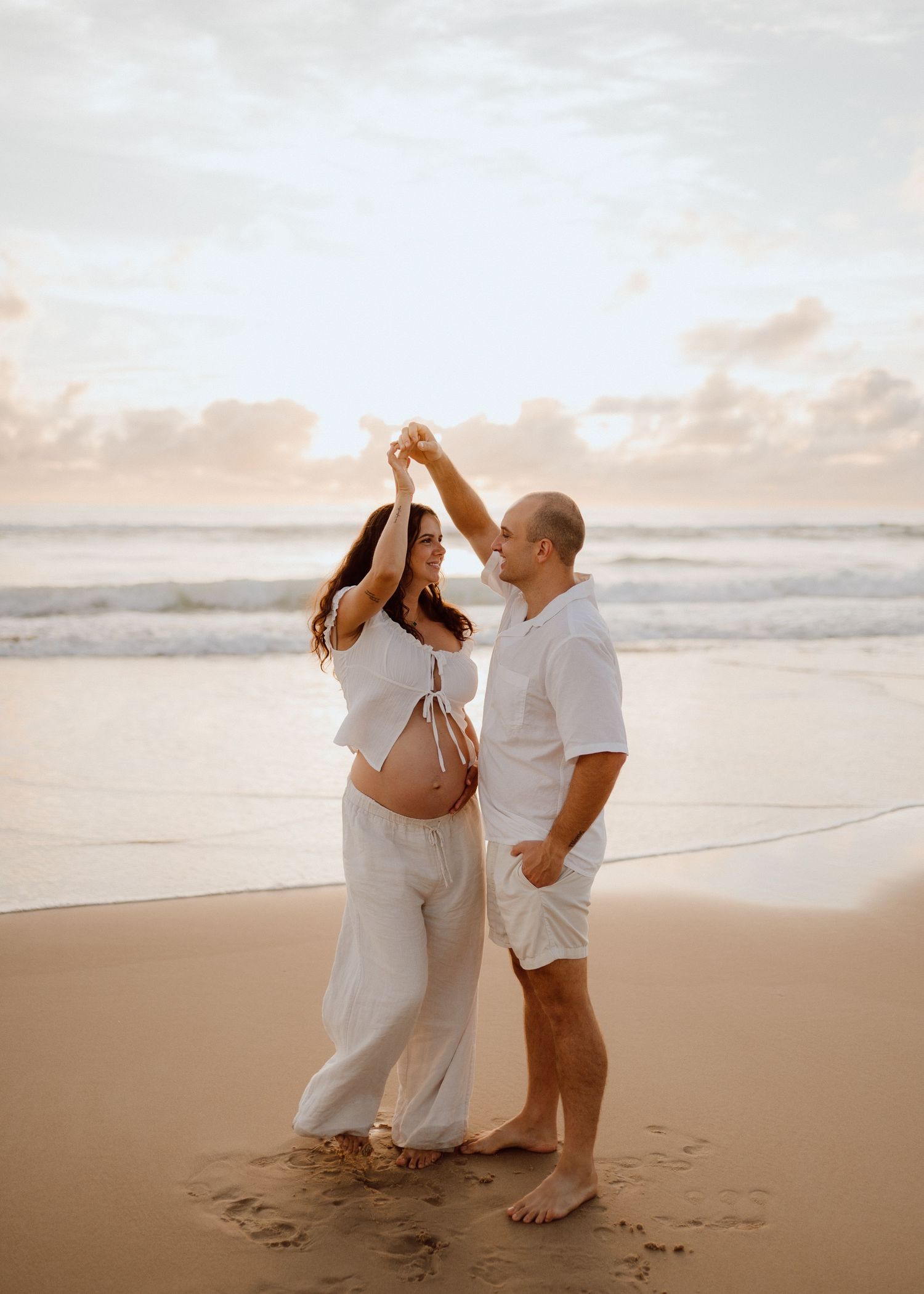 A couple in white outfits dance together on a beach at sunset with ocean waves in the background.