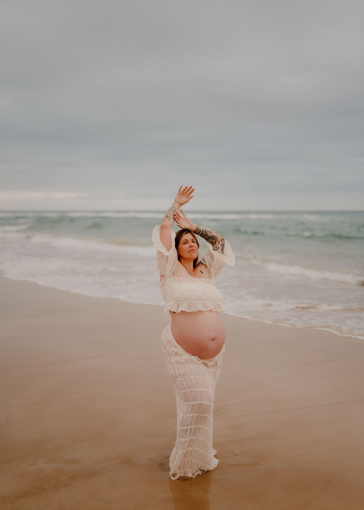 A pregnant woman in a white lace dress poses gracefully on a misty beach at sunset.
