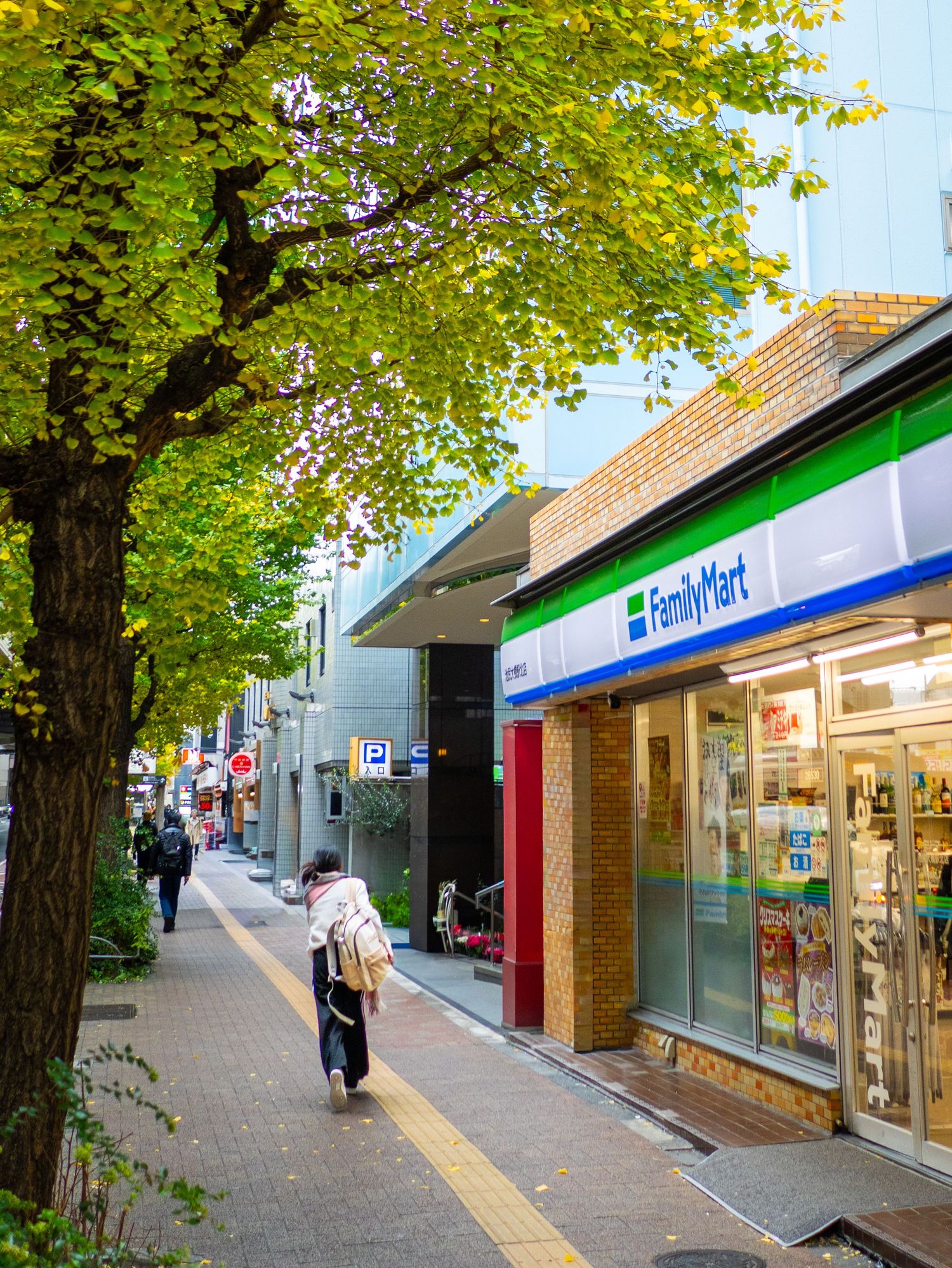 A row of retail storefronts along a tree-lined sidewalk featuring a Family Mart convenience store and other shops.