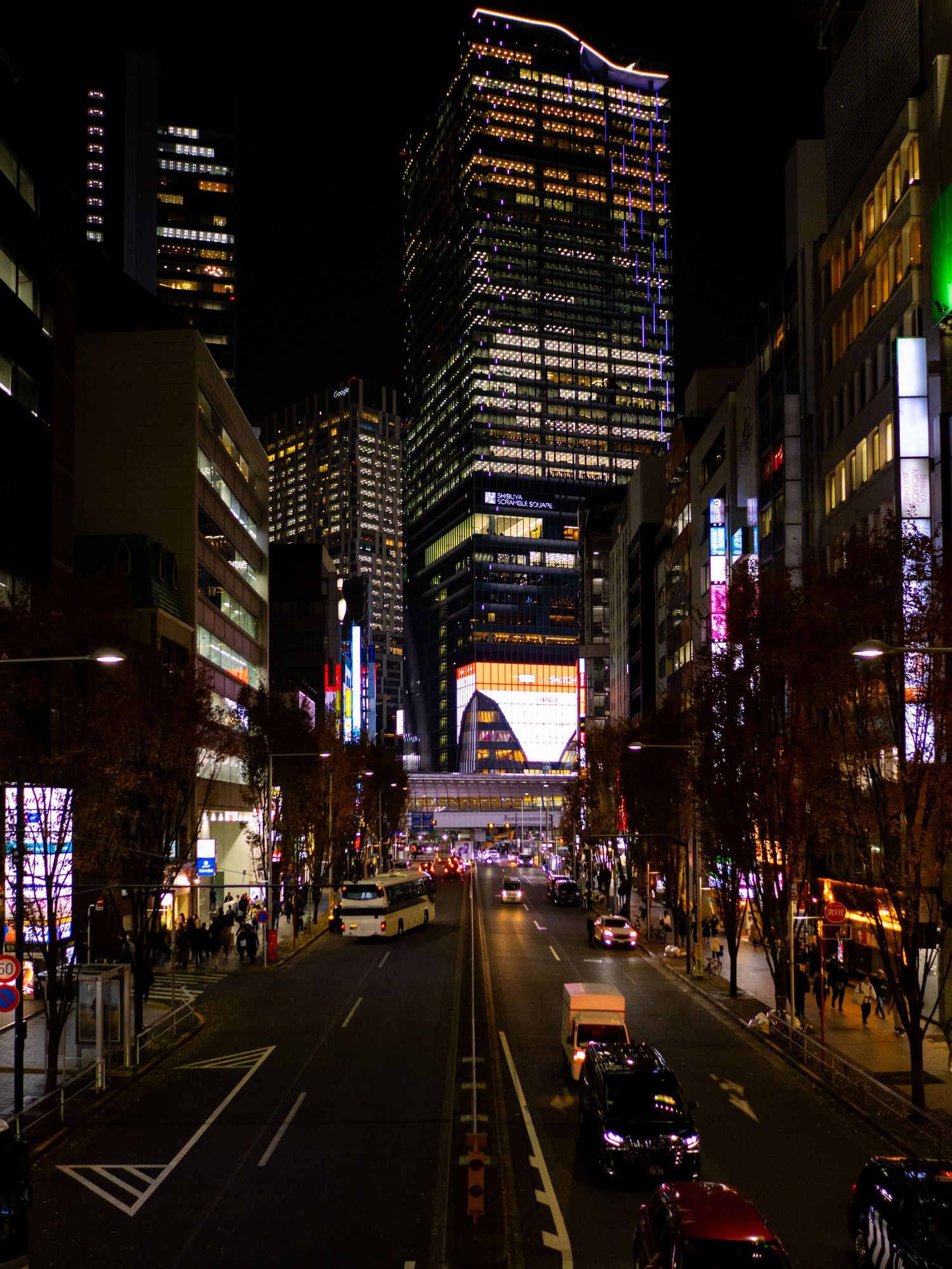 Dark urban street scene with illuminated skyscrapers and neon signs at night.
