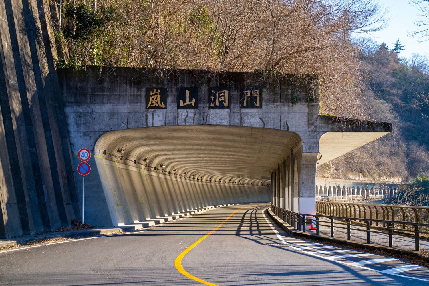 Curved concrete tunnel with repetitive support beams and yellow road line markings winding through a mountainous area.