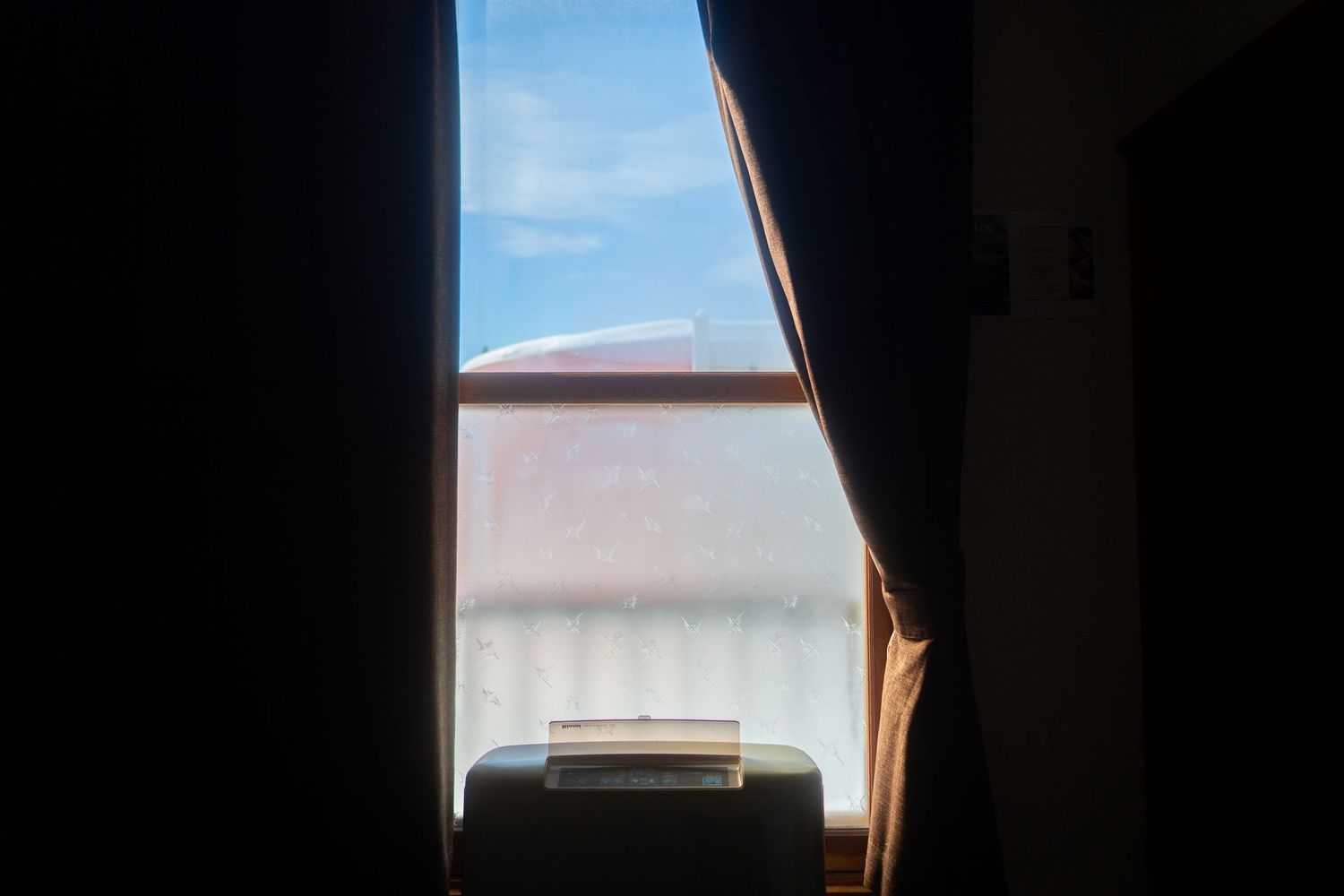 View through a dark curtained window showing a bright blue sky and white building exterior in daylight.