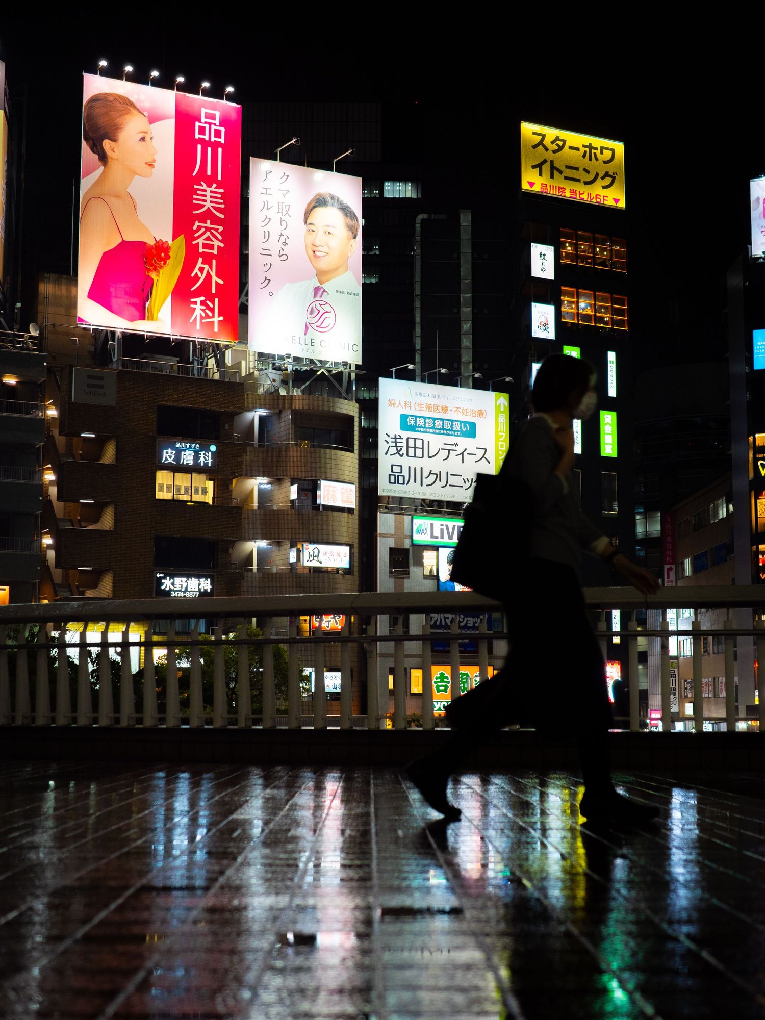 Silhouette of a person walking at night in a rainy Japanese city with colorful neon billboard signs reflecting on wet pavement.
