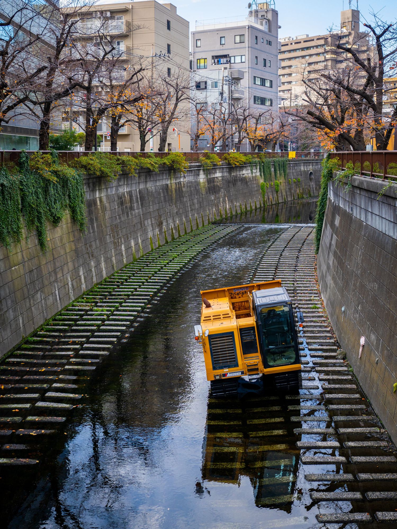 Yellow maintenance vehicle in curved concrete canal with apartment buildings and bare trees along urban waterway in Japan.