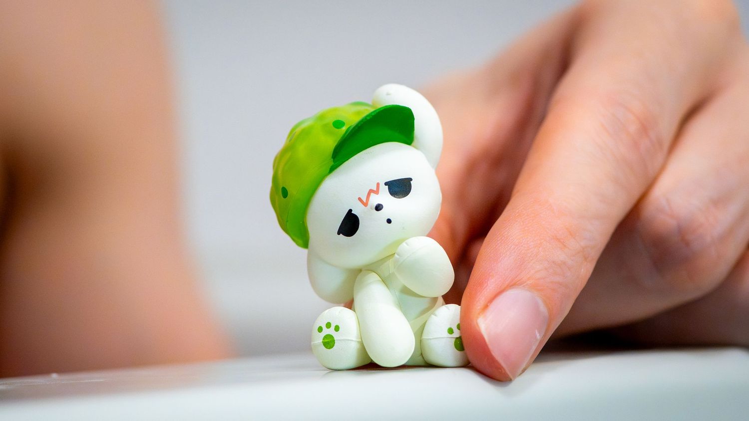 Small white plush toy wearing a green beret cap being touched by a finger on white surface.