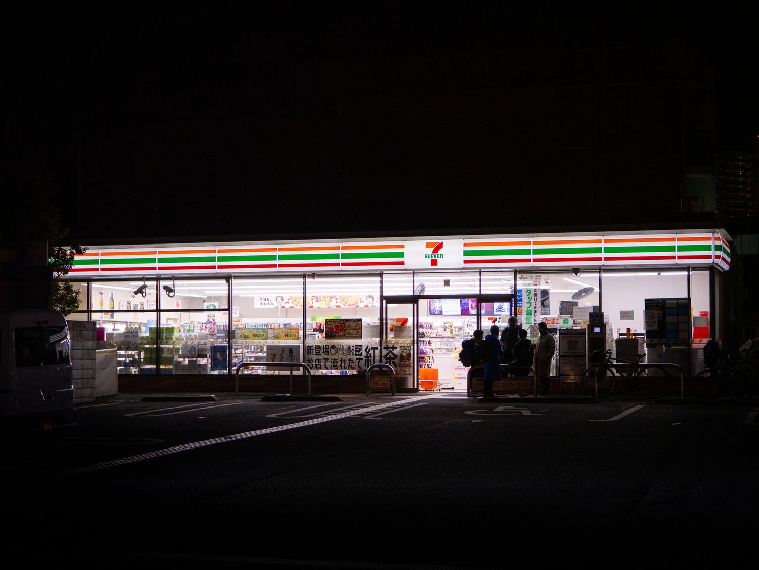 Dark silhouette of buildings with a glowing convenience store sign visible in the distance.