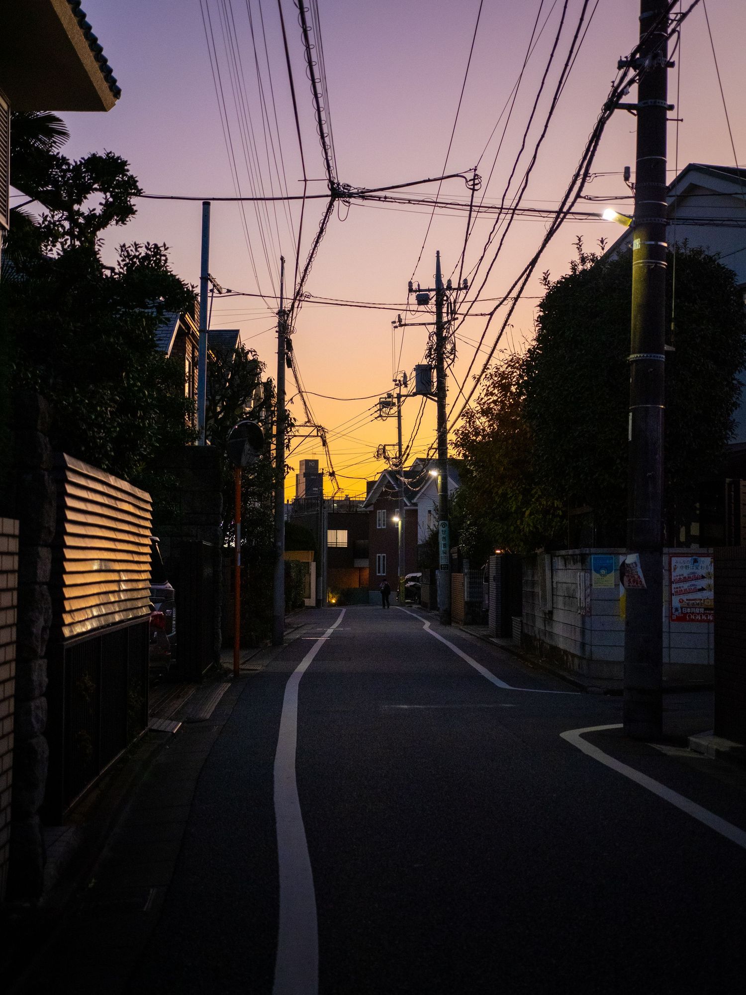 Quiet residential street at dusk with power lines silhouetted against a warm orange and purple sunset sky.