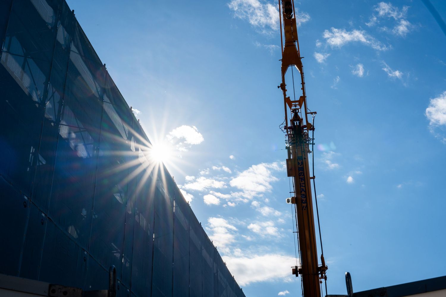 Construction crane silhouetted against bright blue sky with sun flare and scattered white clouds.