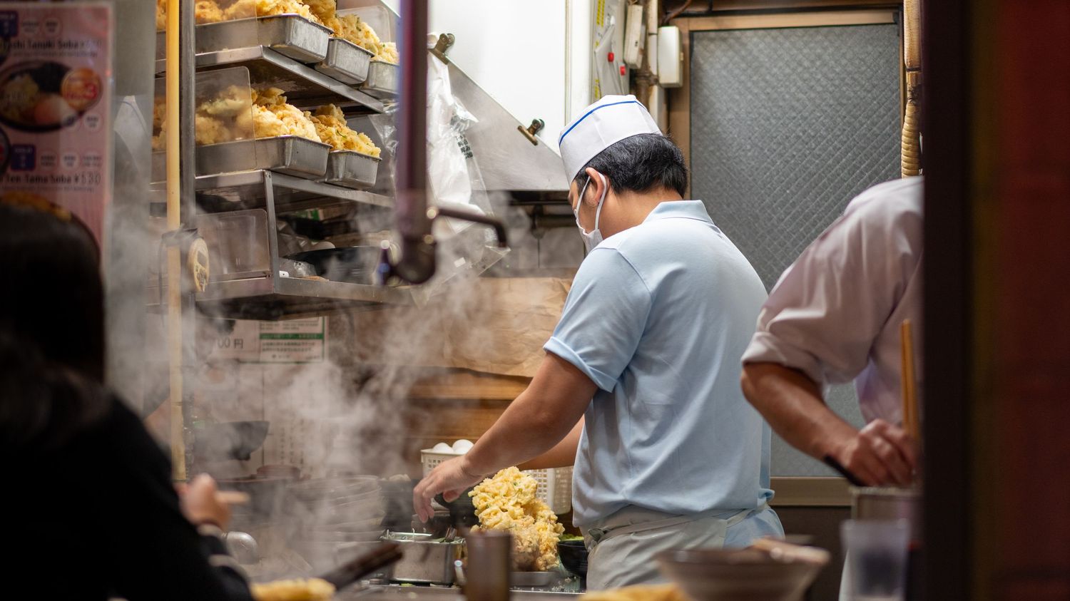 Chef cooking in a steamy restaurant kitchen with food displayed on shelves.