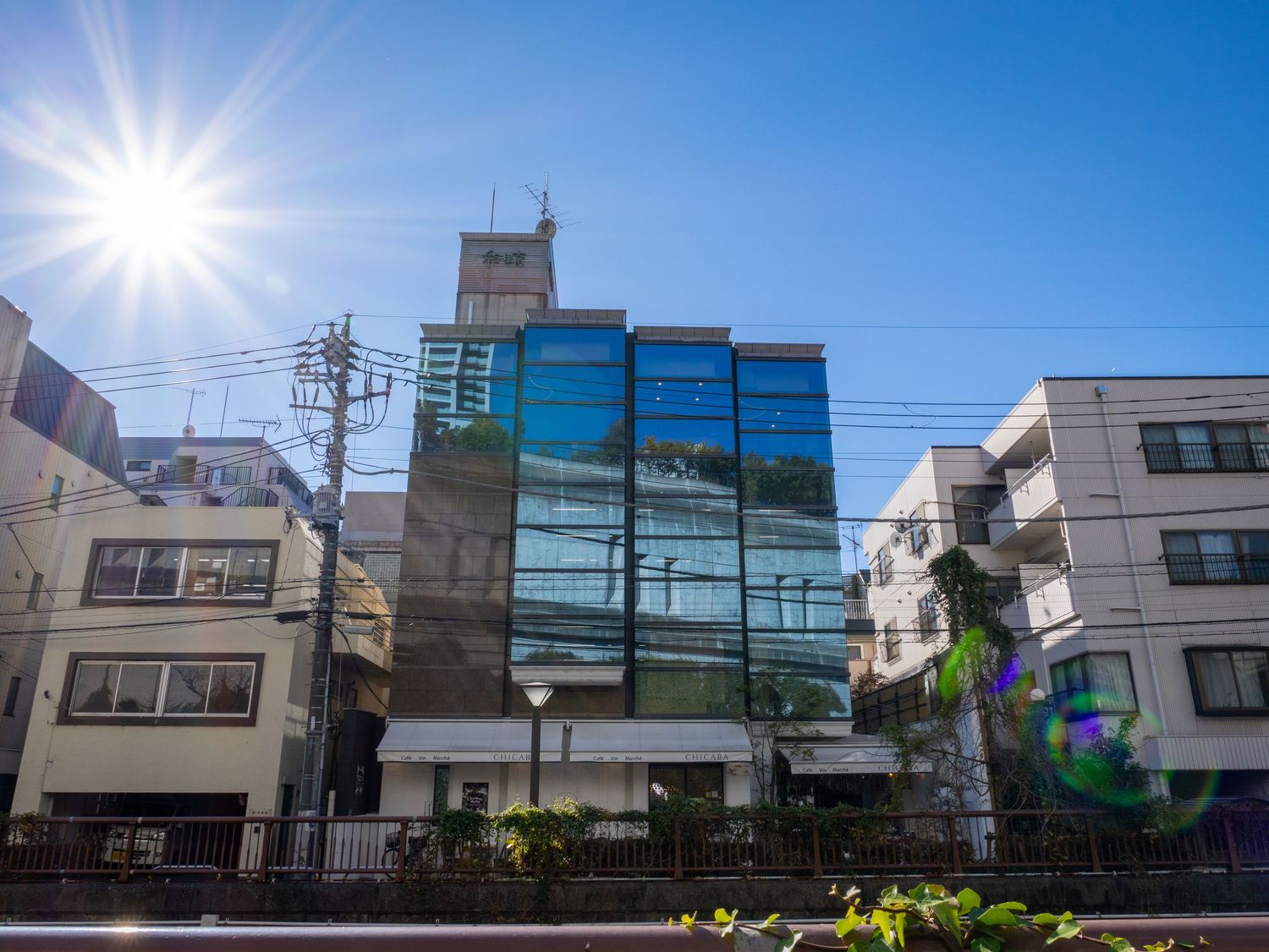 Sun flare shines brightly over modern urban buildings and power lines against a vibrant blue sky.