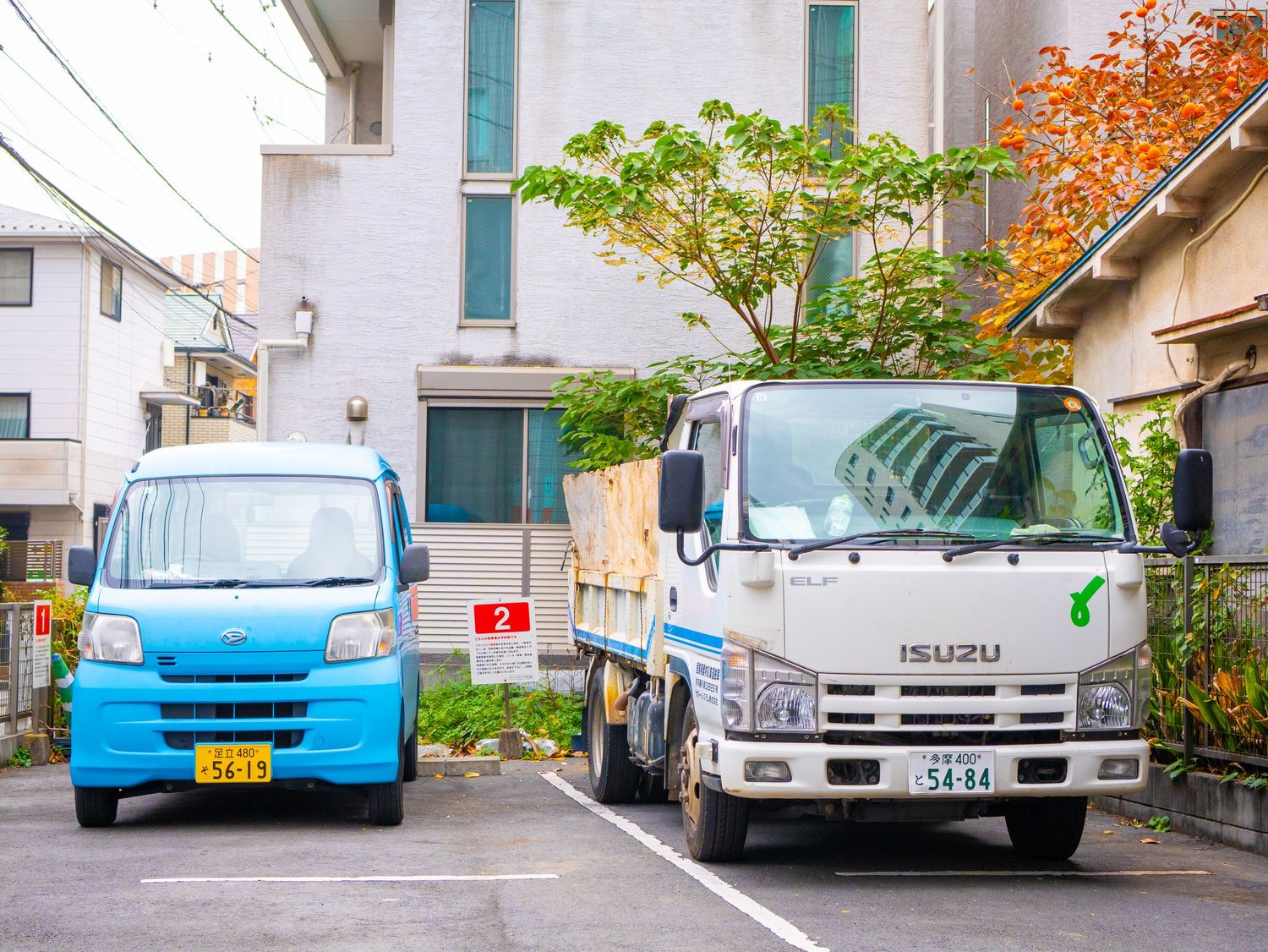 A row of parked cars in front of residential buildings with autumn foliage visible in the background.