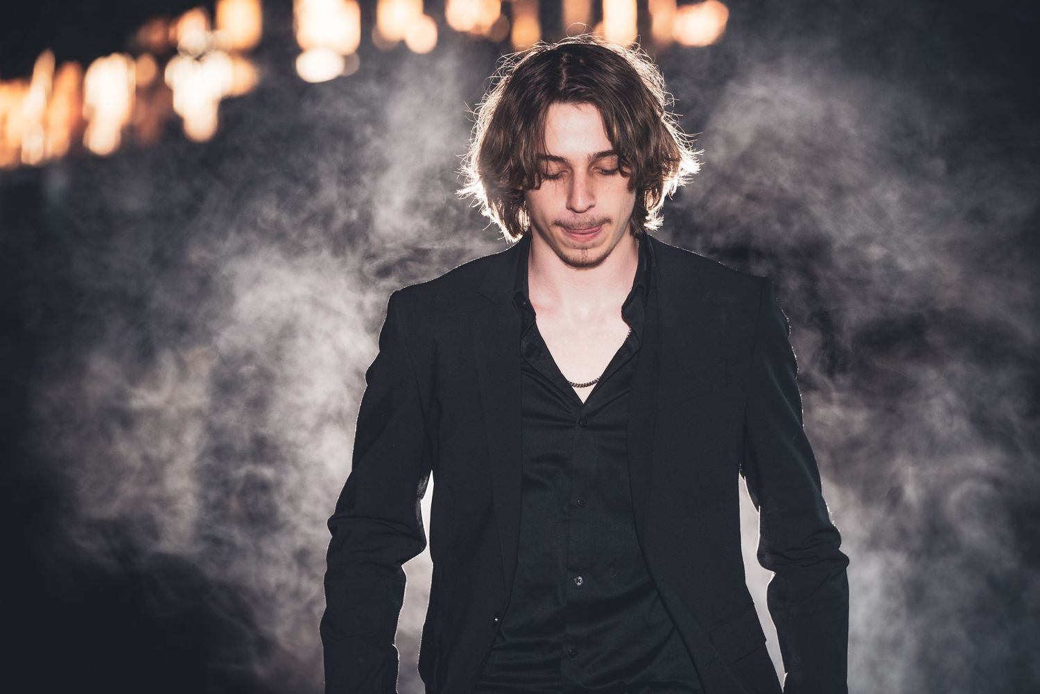 Artistic portrait of someone in a black suit against a smoky dark background.