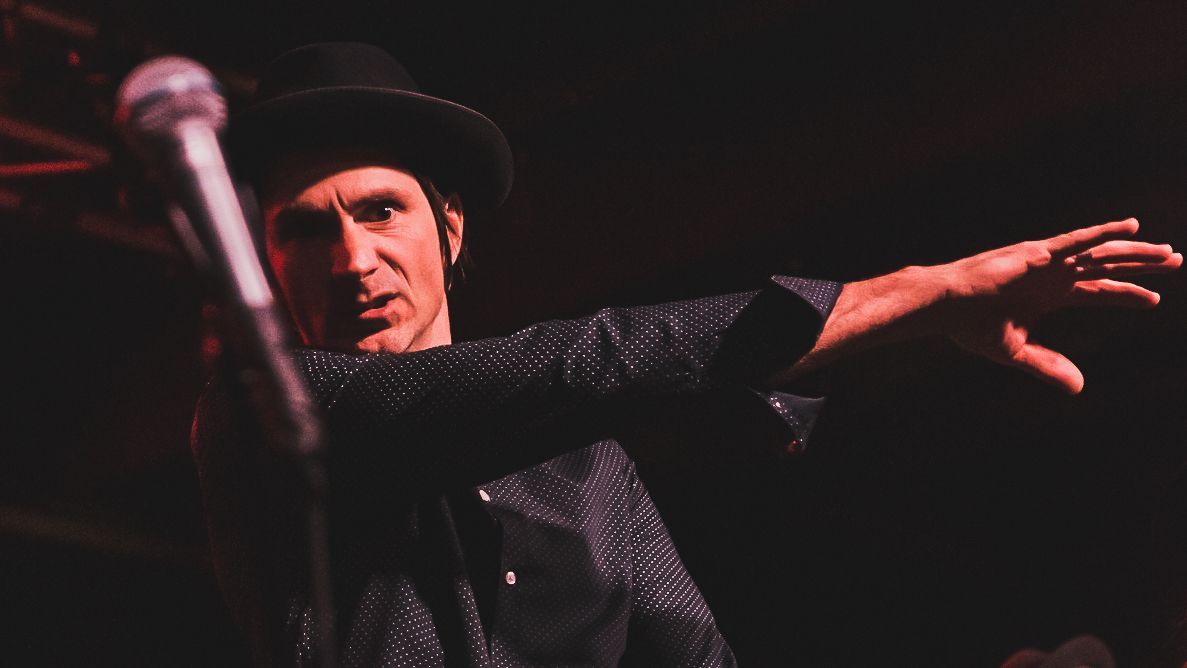 A performer in a black hat gestures dramatically while singing into a microphone on a darkly lit stage.