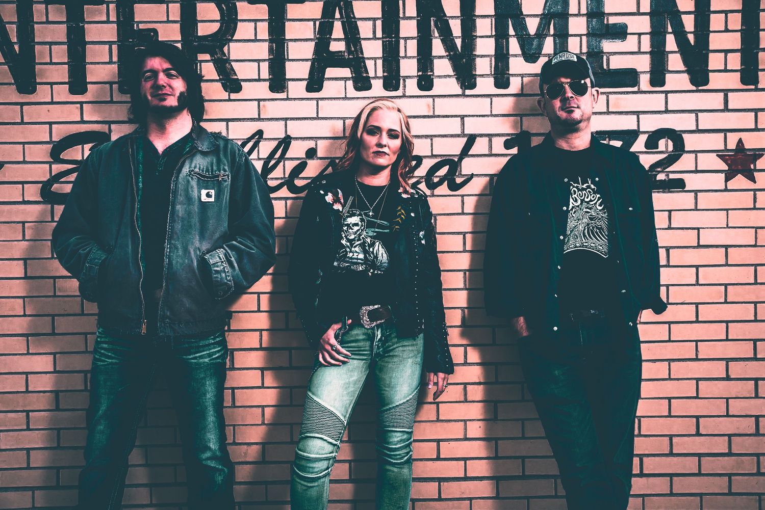 Three musicians pose against a brick wall with Entertainment signage.