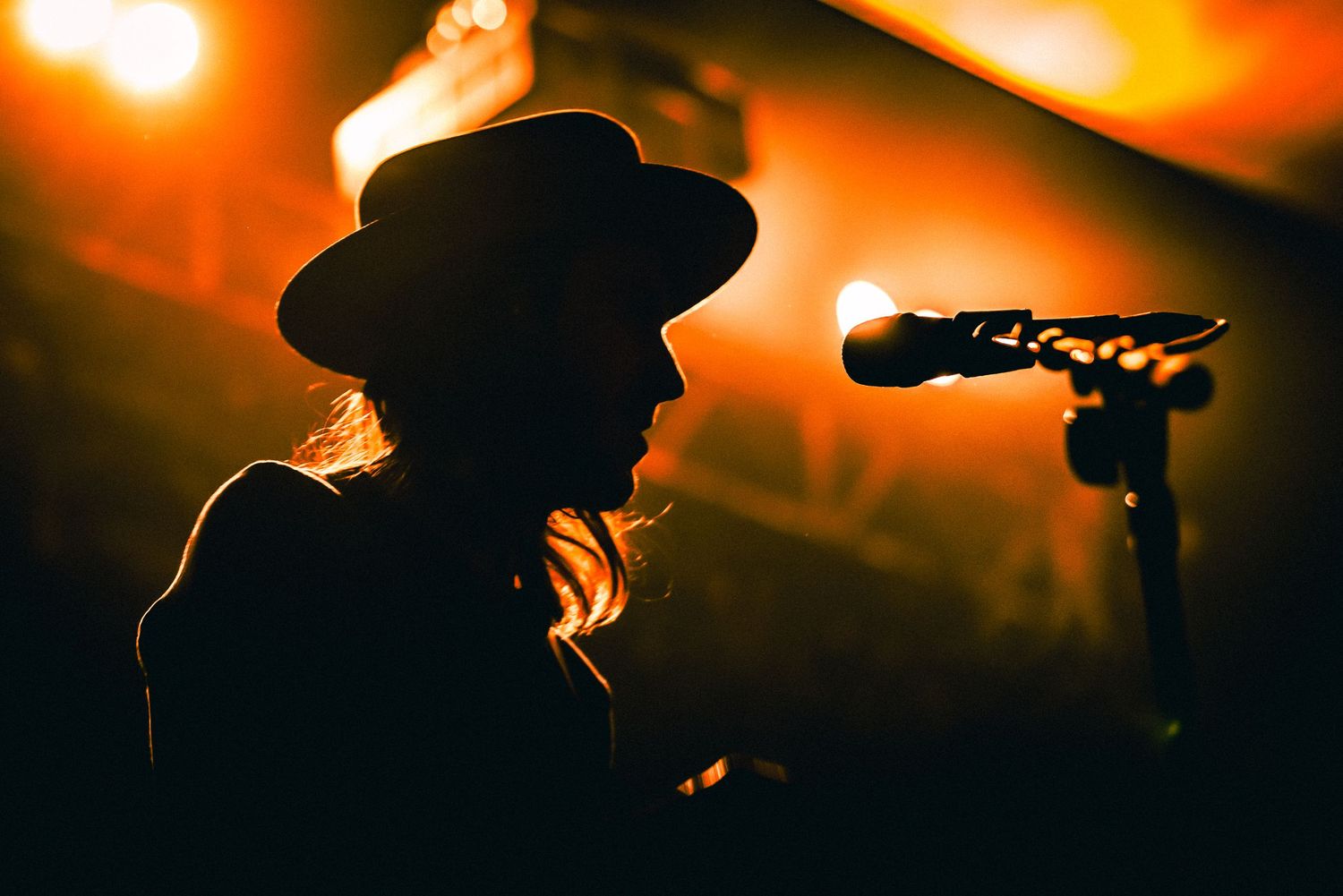 A silhouetted figure in a cowboy hat stands next to a microphone in warm orange stage lighting.
