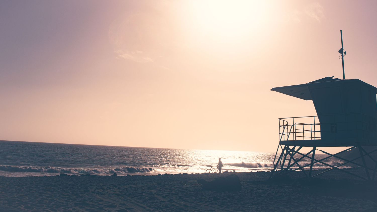 Silhouette of a lifeguard tower against a sunset beach backdrop with sun reflecting on ocean waves.
