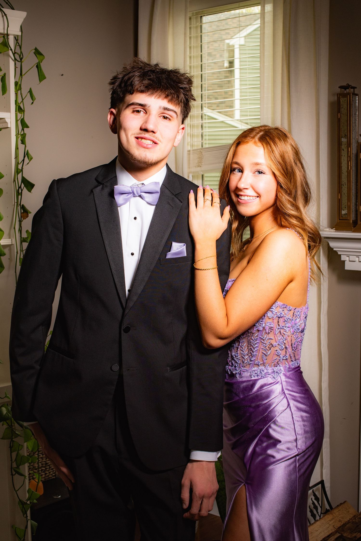 Formal couple posing  for senior photos in Plainfield IL in purple dress and black tuxedo by window with indoor plants.