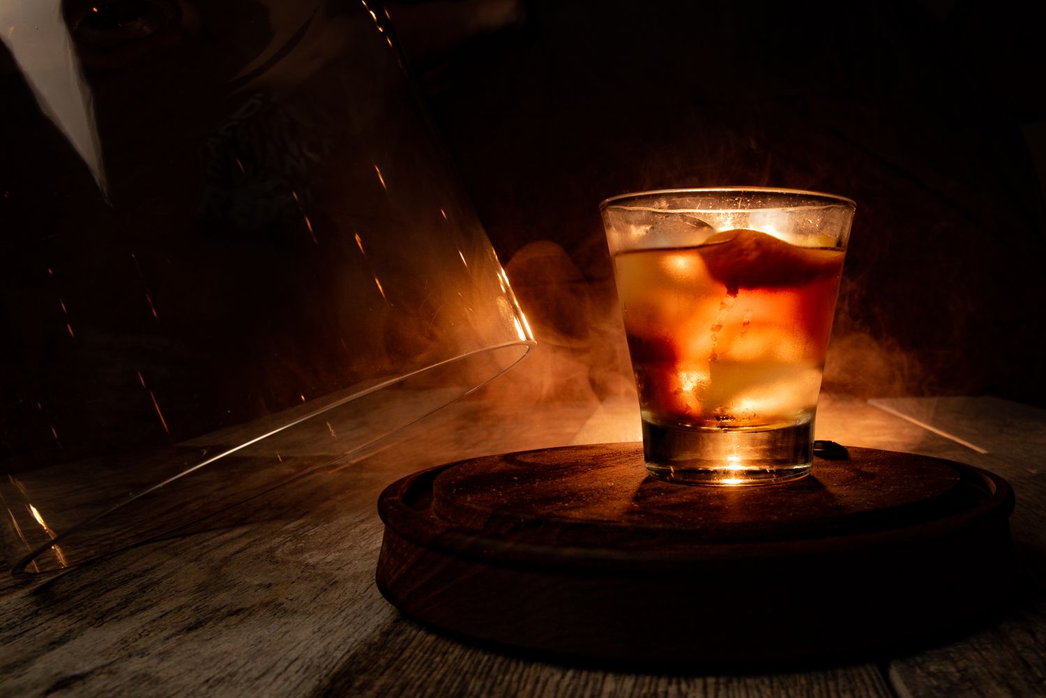 A glass of whiskey with ice sits on a rustic wooden surface illuminated by warm, dramatic lighting.