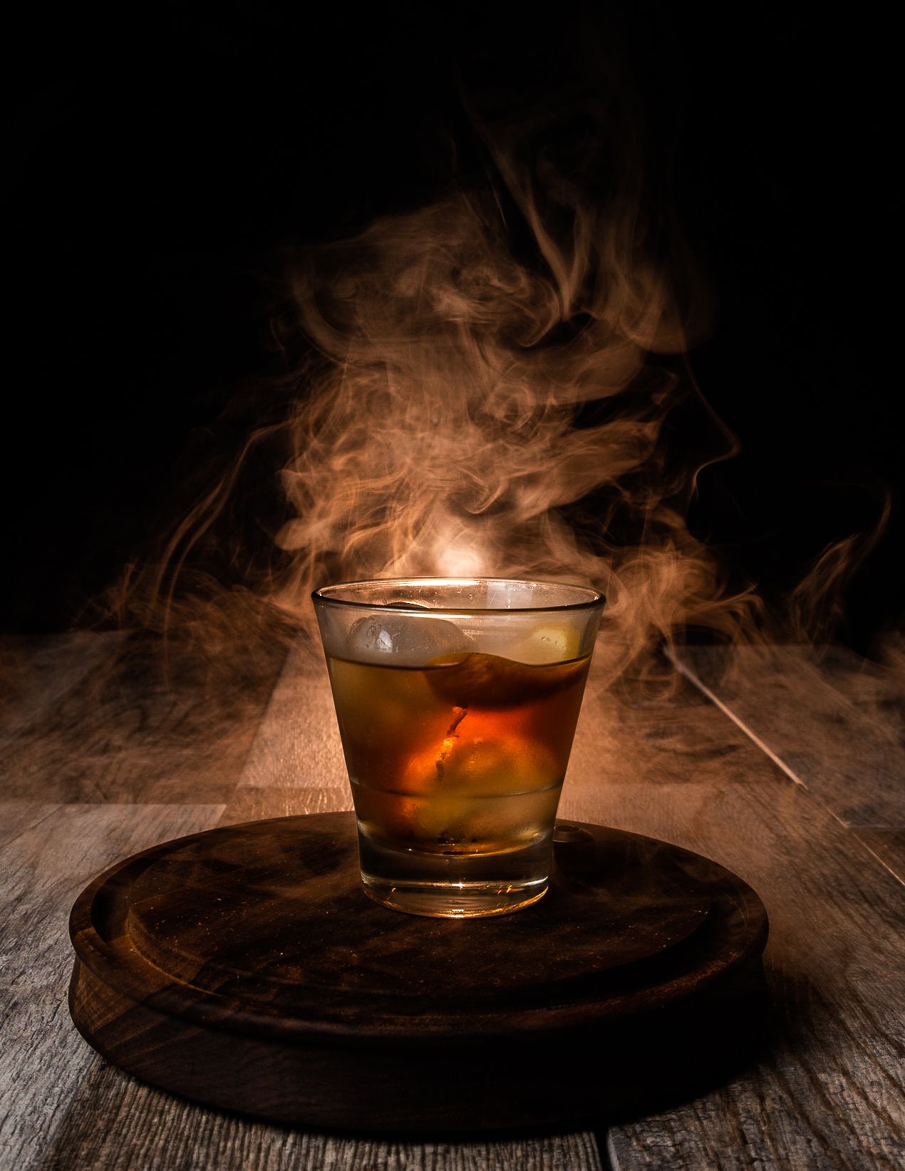 A glass of whiskey on a wooden surface creates dramatic smoke effects in a dark moody setting.