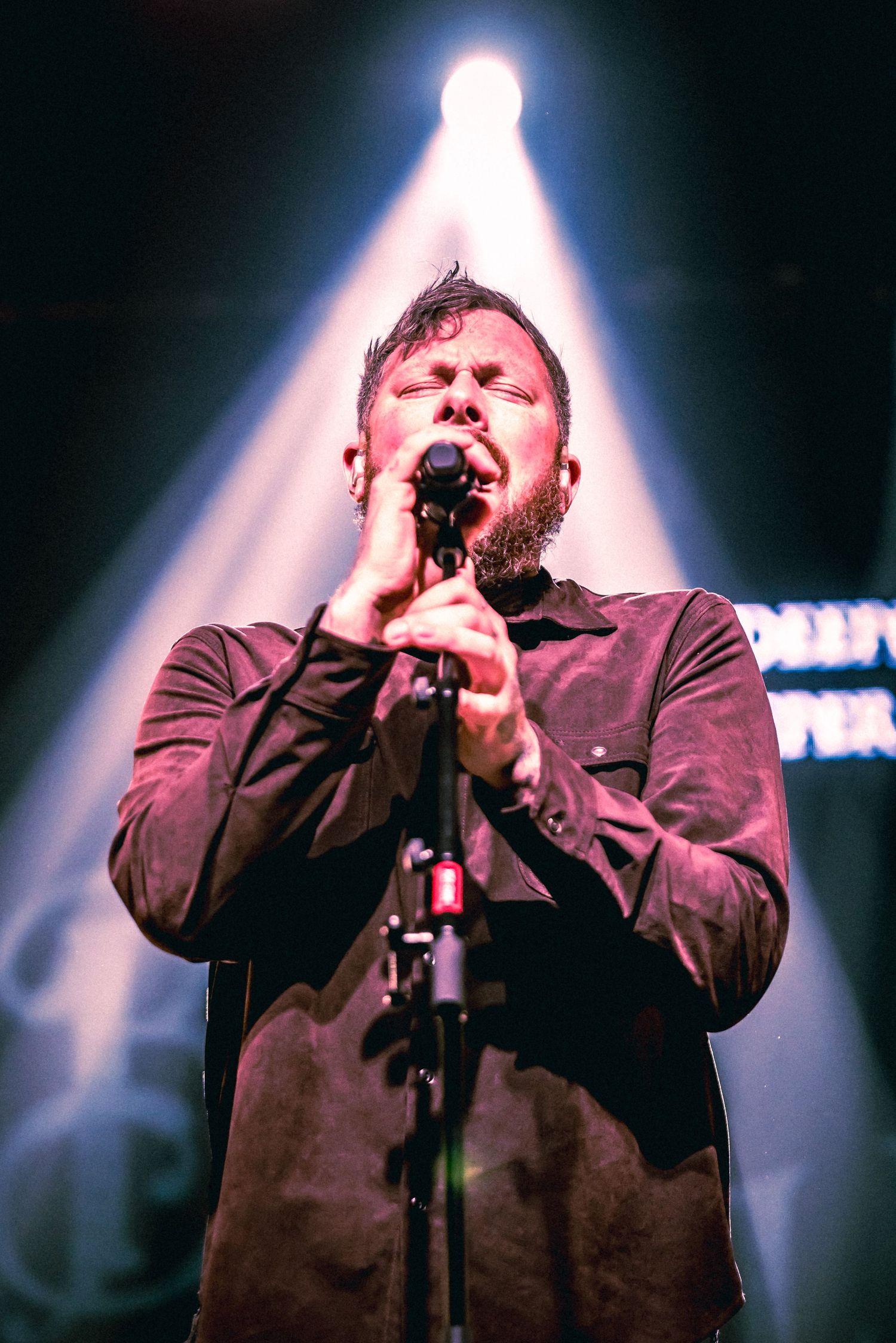 A performer in a dark jacket sings passionately on stage with dramatic backlighting creating a silhouette effect.