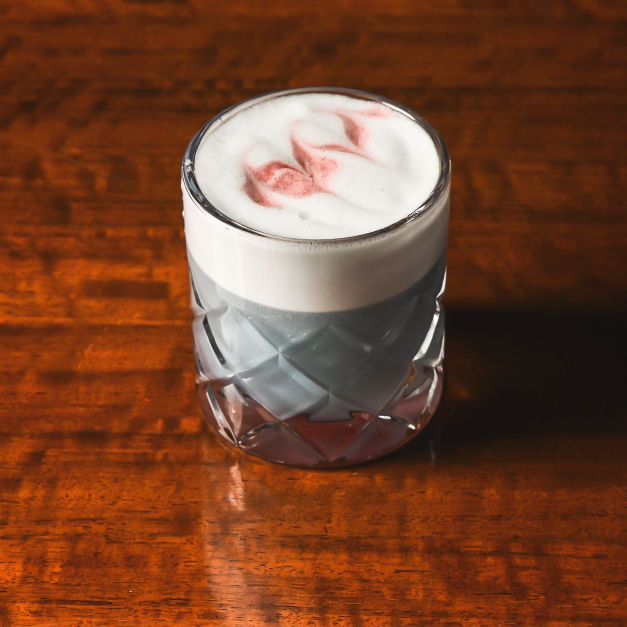 Craft cocktail with white foam top in a diamond-patterned glass on wooden surface.