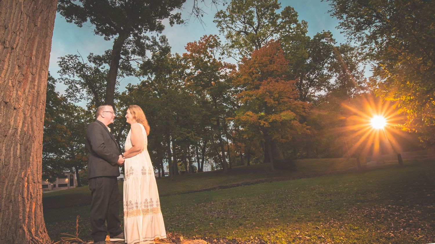 Panoramic sunset view with a bride and a groom in a park with trees and walking path in autumn.