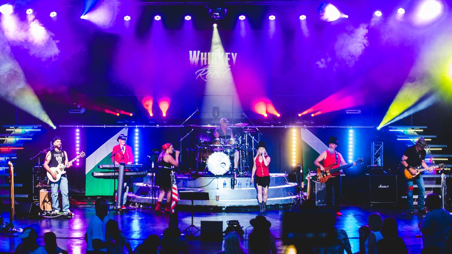 A full band performs on stage with colorful purple and blue lighting and Whiskey River branding.